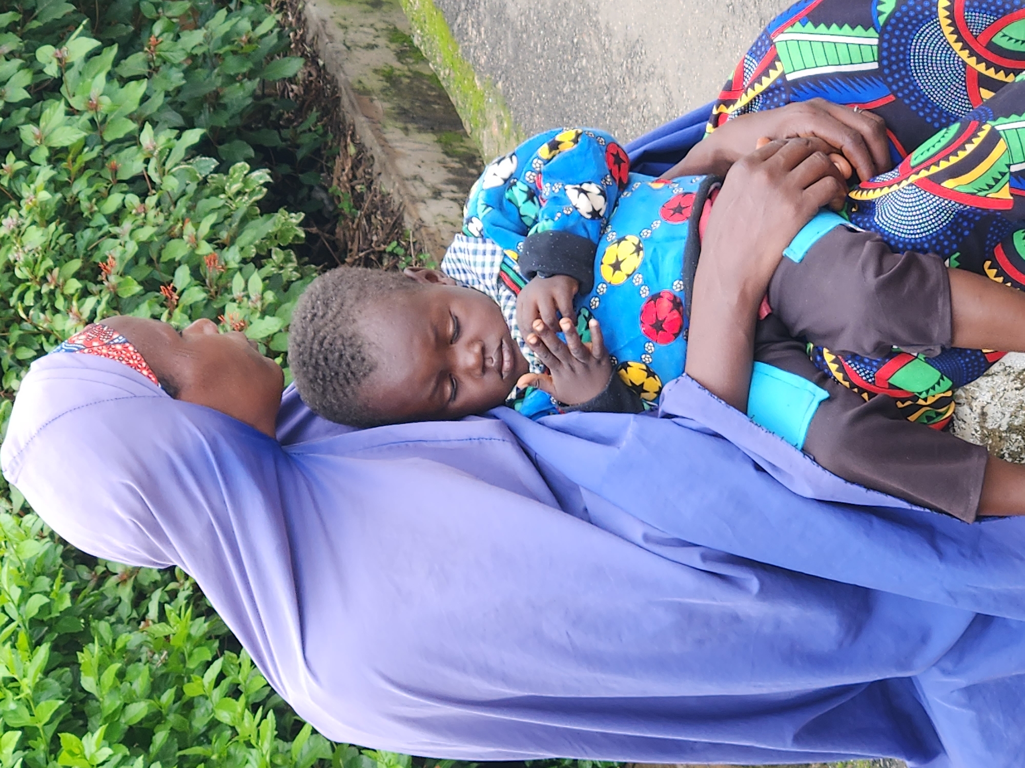 A person wearing a purple hijab and a colorful patterned skirt sits outdoors, holding a sleeping infant. The person's head is turned to the right, with their face partially obscured. The infant, dressed in a blue jacket with a pattern of red, yellow, and white balls, is nestled against the person. Their hands are clasped together, and their eyes are closed. The background consists of lush green foliage and a paved pathway with a concrete border. The scene appears to be in a natural or park-like setting, possibly within the city of Jos, Nigeria, given the presence of vibrant vegetation and local attire.
