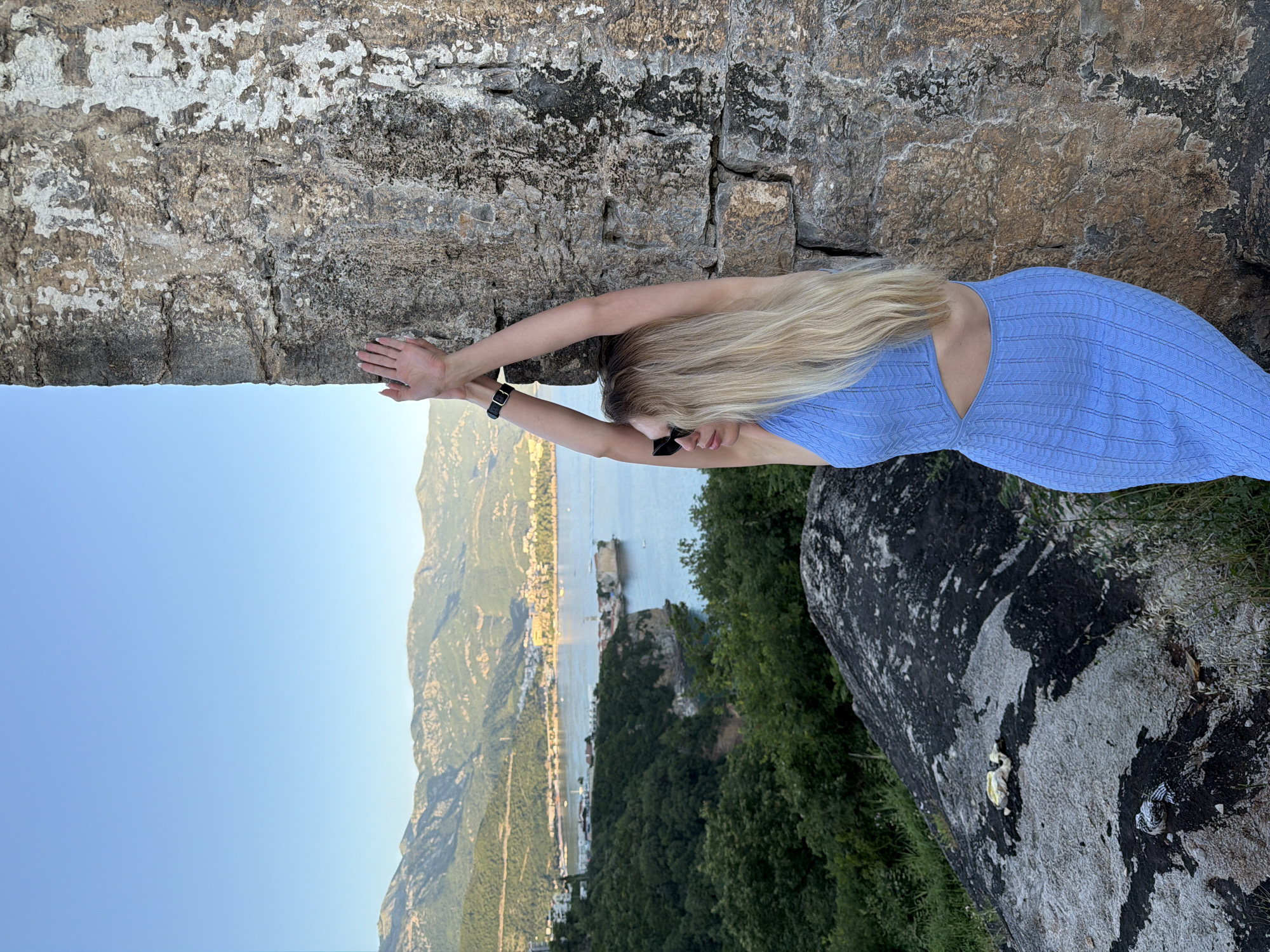 The image captures a woman leaning against a weathered stone wall, overlooking the coastal city of Budva, Montenegro. The woman, with long blonde hair, is wearing a light blue knit dress with cut-outs on the sides and black sunglasses. Her arms are extended upwards, with her hands resting against the textured, grey and white stone wall. A black watch is visible on her left wrist. The background features a wide bay with several boats on the water, bordered by the distinct Old Town of Budva situated on a peninsula, and numerous buildings stretching along the coastline and up into lush green, rocky mountains. The sky above is clear and light blue. In the immediate foreground, a large, dark grey and black rock formation is visible.