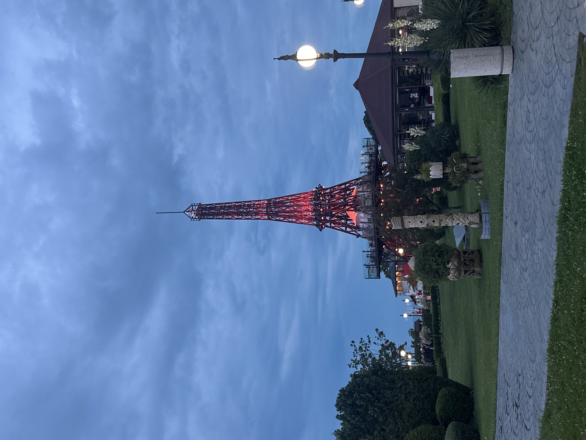 This image depicts a nighttime view of the replica Eiffel Tower at the Sea Garden in Varna, Bulgaria. The tower is illuminated with red lights, casting a glow against the darkening, cloudy sky. In the foreground, a paved walkway leads through a manicured green lawn with sculpted bushes and decorative planters. A lamppost with a lit globe stands to the right of the tower, and a restaurant with a dark-tiled roof is visible behind it. A group of people are gathered in the middle ground, near a line of lampposts. The overall impression is of an outdoor entertainment or dining area, with the iconic tower serving as a prominent landmark.