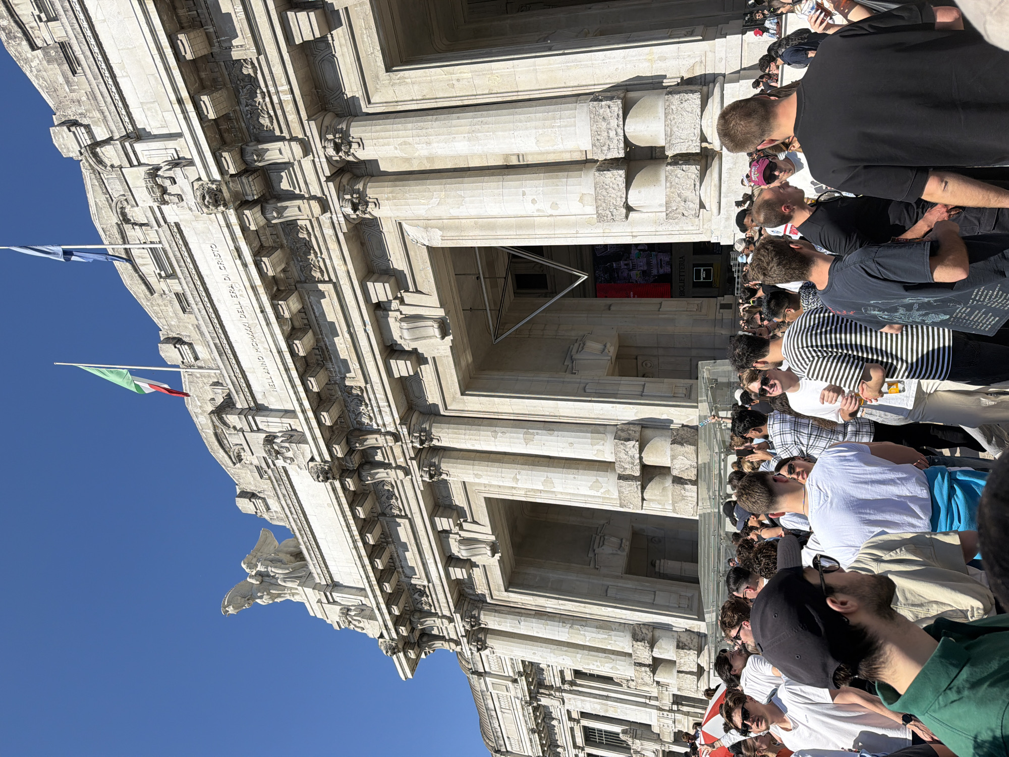 The image shows a crowded scene in front of the Milan Central Station in Italy. The neo-classical facade of the station is visible, including architectural details like columns, cornices, statues, and inscriptions. People are standing close together, likely tourists or travelers, looking at the station, with the Italian and other flags visible. The weather seems sunny, with clear blue skies visible in the background.