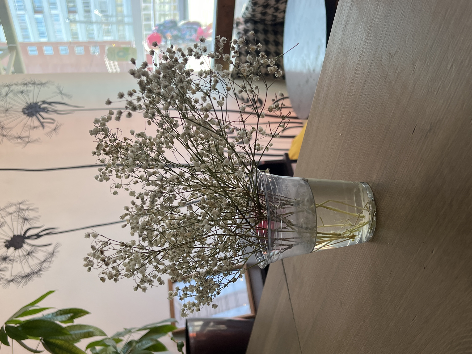 A vase of Baby's Breath flowers sits on a wooden table. In the background, a wall is visible with a dandelion mural. Through a window, city buildings are seen. While the prompt suggests the location is Berezniki, Russia, there is no way to confirm the location from the image alone.