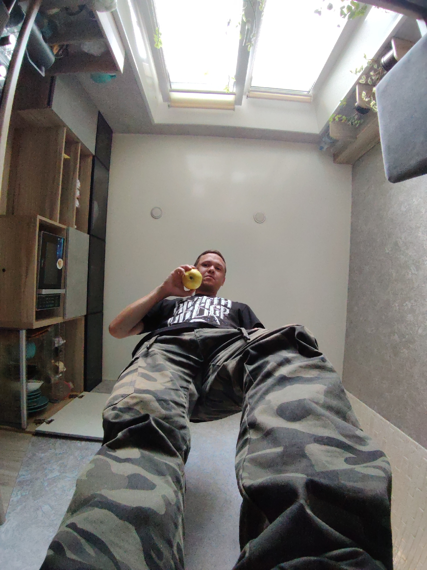 A man in camouflage pants and a black t-shirt holds a yellow apple in his right hand and looks directly at the camera. The image appears to be taken from a low angle within an indoor setting. To the left of the man, there is a wooden shelving unit with a microwave and various items on the shelves. The wall behind the man is white, and the ceiling features two skylights with sunlight filtering through. The floor is a light gray tiled surface. The overall environment suggests a domestic or residential space.