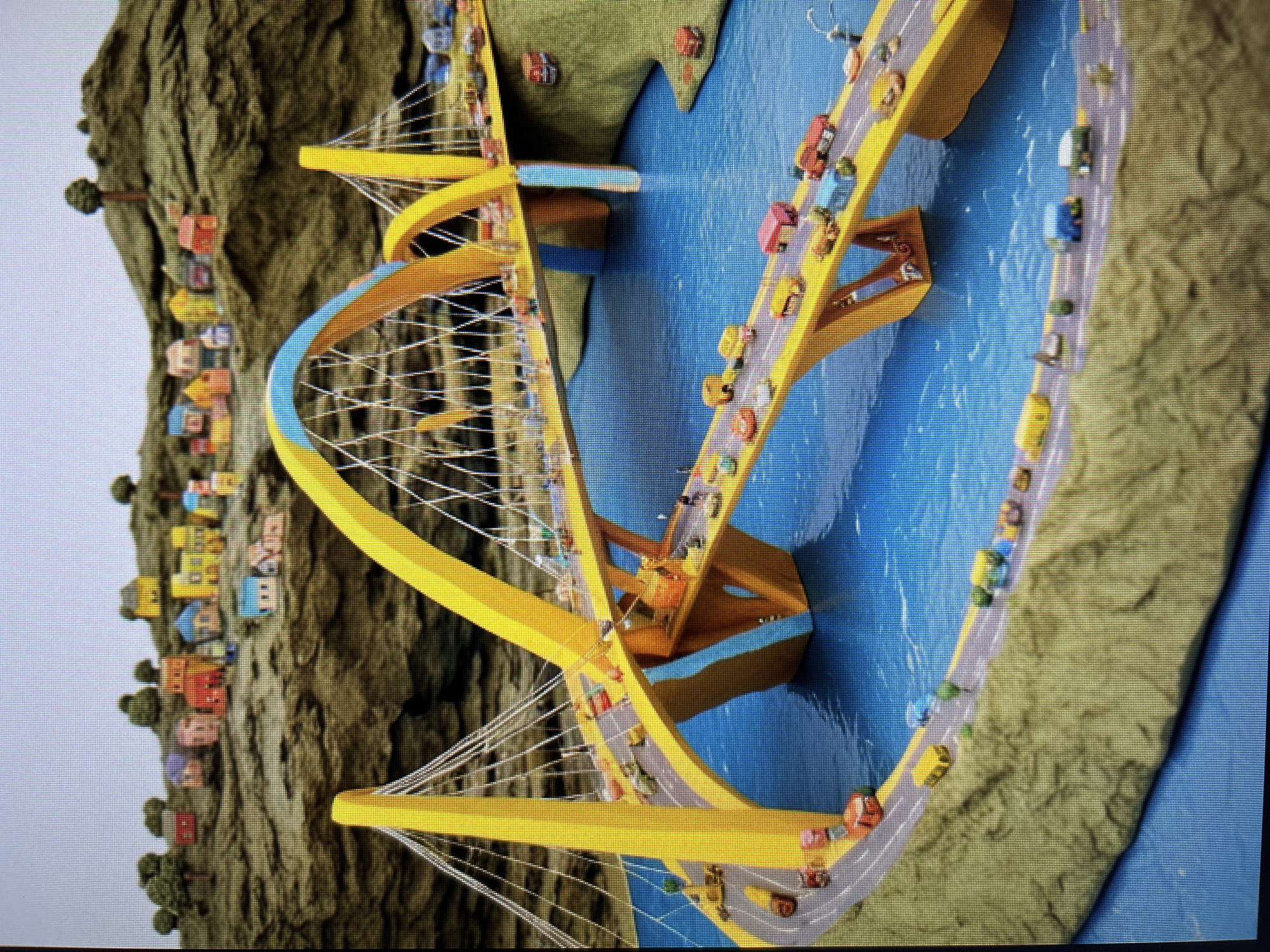 The image depicts a miniature diorama of a brightly colored city scene. A large, yellow suspension bridge with blue accents spans across bright blue water. The bridge is populated with numerous small, toy cars of various colors, indicating traffic. The shoreline of the diorama is represented by a textured, green-brown material, resembling rolling hills. On these hills, a cluster of small, toy houses with colorful roofs are arranged, resembling a village. The sky is a light gray. The overall impression is that of a whimsical, toy-like representation of an urban landscape.