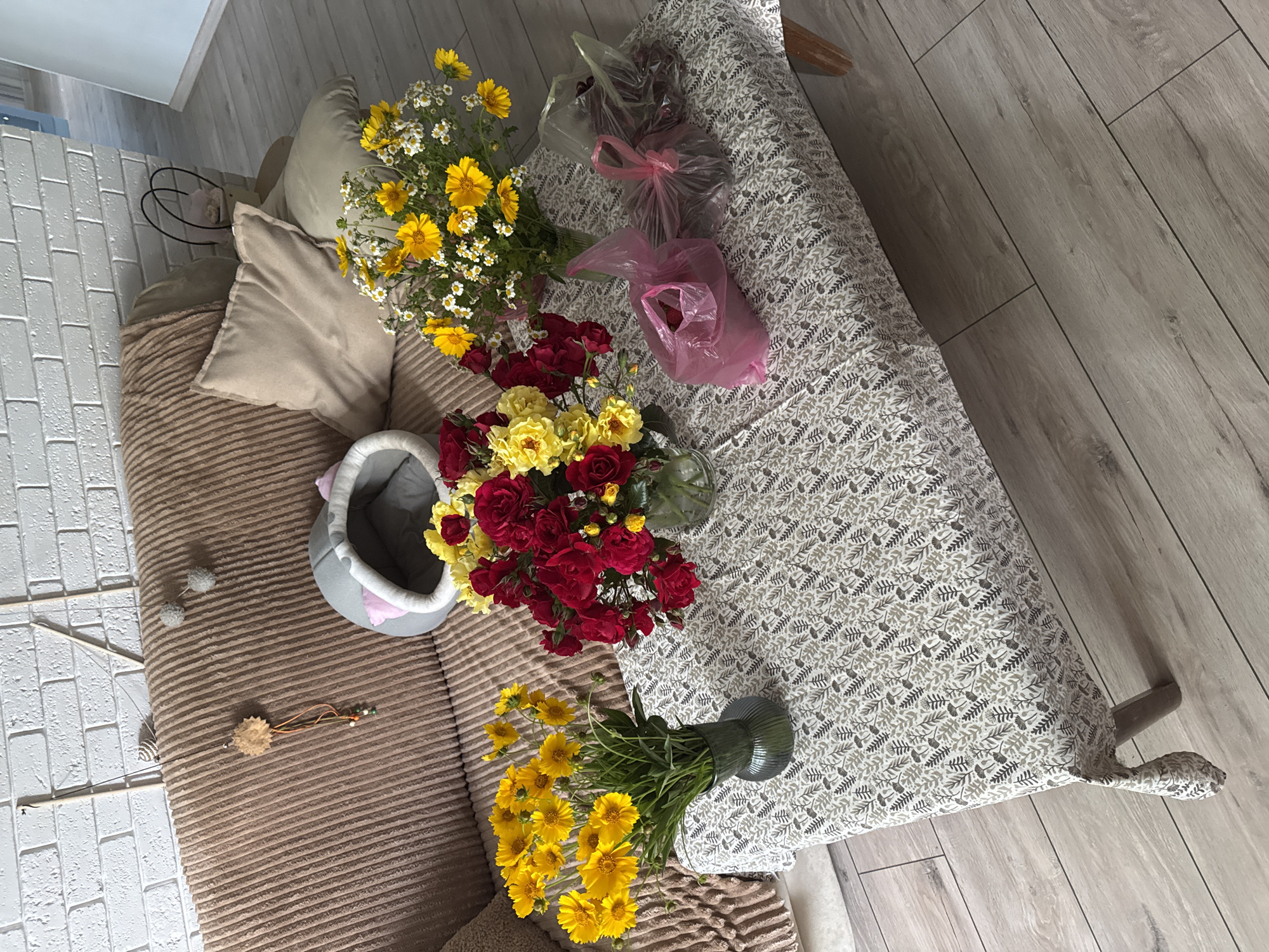 A high-angle medium shot shows a couch and a table covered with a patterned tablecloth. On the couch, a light brown blanket is draped over the back and arms, with two light beige throw pillows and a gray cat bed with pink trim. A wooden cat toy with feathers and beads hangs from a stick on the wall. On the table, there are three bouquets of flowers in vases, two in pink plastic bags, and a third in a clear plastic bag filled with red berries. The floor is made of light gray wood-look laminate. The scene is indoors, in what appears to be a living room.
