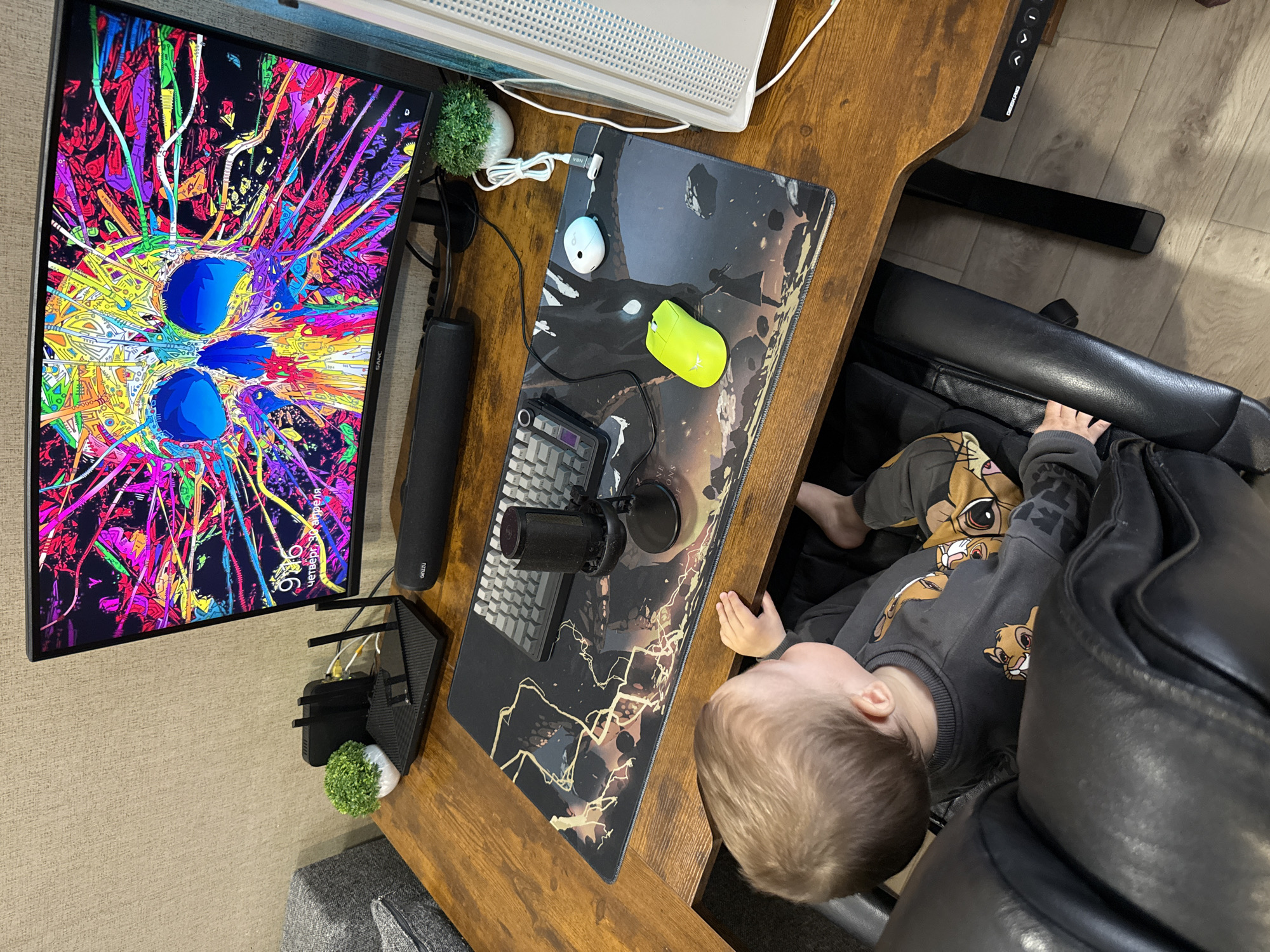 An overhead shot captures a young child interacting with a computer desk setup in an indoor setting, identified as Rusanovo, Russia.
**Subjects:** The primary subject is a young child with light hair, seen from above, wearing a gray long-sleeved shirt patterned with multiple "Simba" characters from The Lion King. The child is positioned next to or in a black leather office chair, with bare feet visible. The second main subject is an L-shaped wooden desk equipped with a computer monitor, keyboard, microphone, and other accessories.
**Objects:** The monitor is oriented vertically, displaying a vibrant, abstract, and highly colorful digital artwork featuring swirling lines and three prominent blue circular shapes. The monitor bezel has the brand name "SANC". A black speaker bar is positioned below the monitor. The wooden desk, which appears to be a standing desk with control buttons ("M1", "M2", up/down arrows) visible on its right edge, holds a large mouse pad with a dark, abstract design, possibly depicting lightning or cracks. On the mouse pad are a neon green computer mouse and a white/light blue mouse, along with a mechanical keyboard and a black microphone on a stand. A small artificial green plant in a white pot, a white cable, and black network equipment (including a router with antennae) are also on the desk. The floor is a light-colored wood, with a gray textured carpet or rug partially visible.
**Activity/Event:** The child appears to be reaching towards the desk or observing its contents, possibly playing near the workspace.
**Notable Details:** The monitor displays the time as "9:16" and the date as "Четверг, 30 апреля" (Thursday, April 30th), suggesting the time of day the photo was taken. The overall setup indicates a home office or gaming station. The child's clothing suggests a casual indoor environment. The image is brightly lit, likely from ambient indoor light.