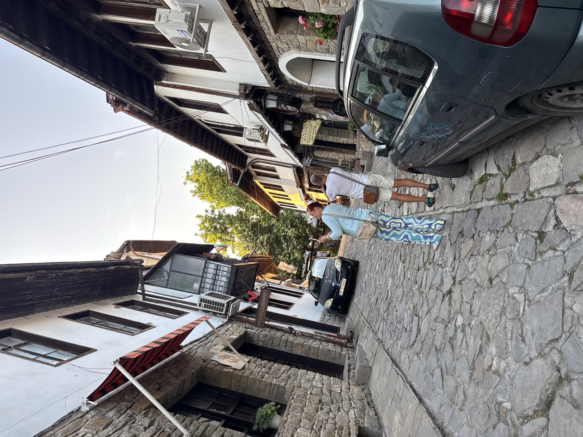 In Veliko Tarnovo, Bulgaria, a narrow, cobblestone street is lined with historic buildings. Two individuals, a woman and a man, stand in the street, with the woman taking a photograph of the man. The man faces away from the camera, looking up the street. In the foreground, a dark teal car is parked on the right side of the street. The buildings feature stone foundations and white stucco walls, with wooden window frames and some featuring awnings. Signs for businesses, including a winery, are visible on the buildings. The sky is clear and bright, suggesting it is daytime.