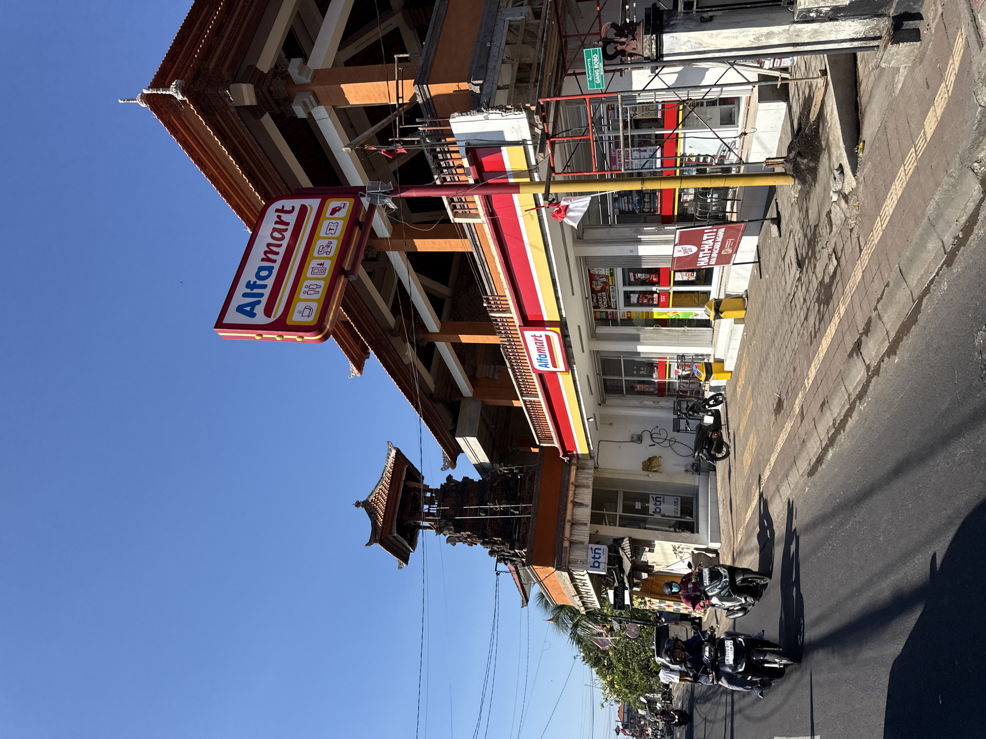 In Denpasar, Indonesia, a street scene unfolds under a clear blue sky. The image is dominated by the facade of an Alfamart convenience store, a prominent red, white, and yellow sign advertising the brand. The building itself features traditional Balinese architectural elements, including ornate roof details and wooden accents, suggesting a local design influence. Scaffolding indicates ongoing construction or renovation on the right side of the building.
In front of the Alfamart, a sidewalk is visible with textured paving for visually impaired pedestrians. Several people are present. Two individuals are riding motorcycles on the street in the foreground; one motorcycle carries two people, both wearing helmets, and the other carries a single rider also wearing a helmet. Other people can be seen walking on the sidewalk and entering or exiting the store. A traditional Balinese temple-like structure with a tiered roof is visible on the left side of the image, adding to the cultural context. Yellow trash bins are placed on the sidewalk. A sign in Indonesian, "GANG BOBO," is mounted on a pole on the right side of the street, indicating a narrow alley or lane. The street is paved and appears to be a main thoroughfare with a moderate amount of traffic. Shadows from overhead wires and buildings are cast across the street and sidewalk.