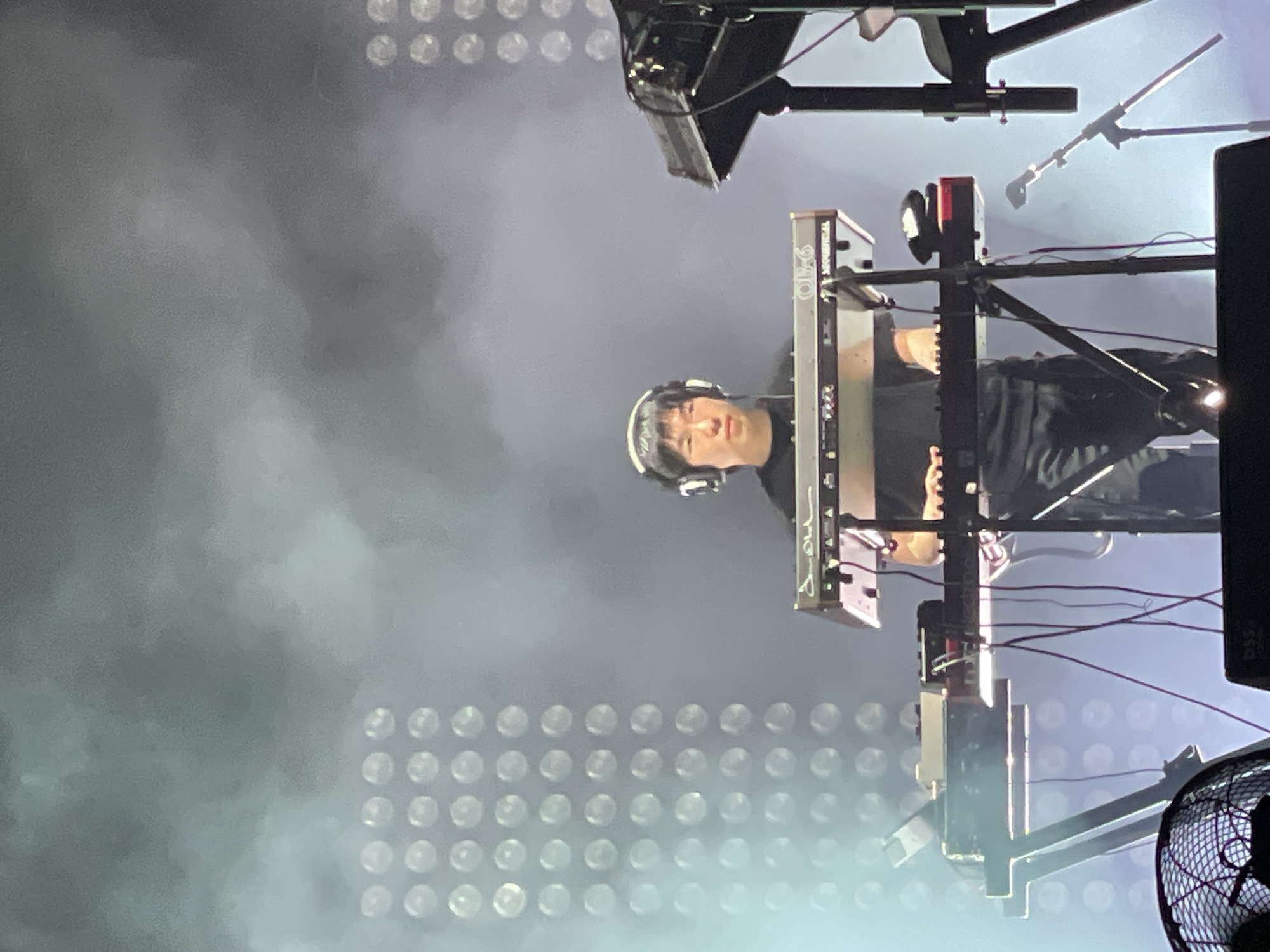 The image appears to show a musician performing on a keyboard synthesizer during a concert. The musician is wearing headphones and dressed in dark clothing. The keyboard is mounted on a stand and surrounded by various pieces of equipment, including a microphone, cables, and a speaker. In the background is a wall of stage lighting that creates a hazy environment. The location may be Salembaran, Indonesia, but this cannot be confirmed from the image itself.