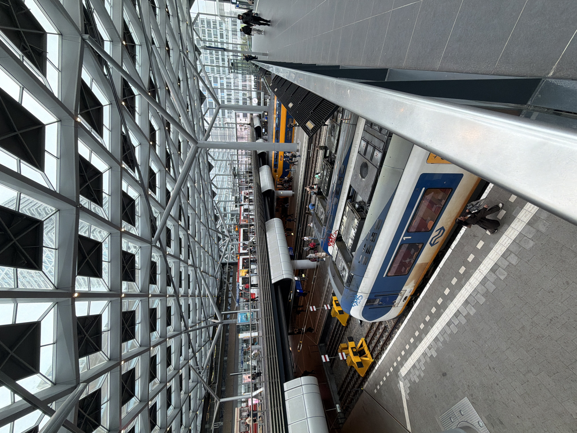 The image depicts a modern train station or terminal from an elevated, diagonal perspective, looking down onto multiple platforms and tracks. A prominent architectural feature, a vast glass and metal grid structure with repeating square and triangular patterns, forms the left side of the frame, possibly part of the roof or a large wall, reflecting ambient light.
Below, two trains are visible. In the foreground on the right, a blue and white train with graffiti (reading "Stas" or similar) on its side and the number "3742" near its front is parked at a platform. A yellow and blue train, recognizable as a Nederlandse Spoorwegen (NS) service, is further back on an adjacent track.
Several people populate the scene. Some individuals are walking on an upper-level concourse visible towards the top-left, while others are on the lower platforms next to the trains. One person is distinctly sitting on the edge of the platform next to the blue and white train in the lower-right corner. The platforms are paved with grey tiles and marked with white dashed and solid lines.
The lighting suggests it is daytime, likely overcast, with a soft, diffused light illuminating the station. Yellow safety barriers are visible on the tracks near the trains. The overall impression is that of a bustling, modern transportation hub.