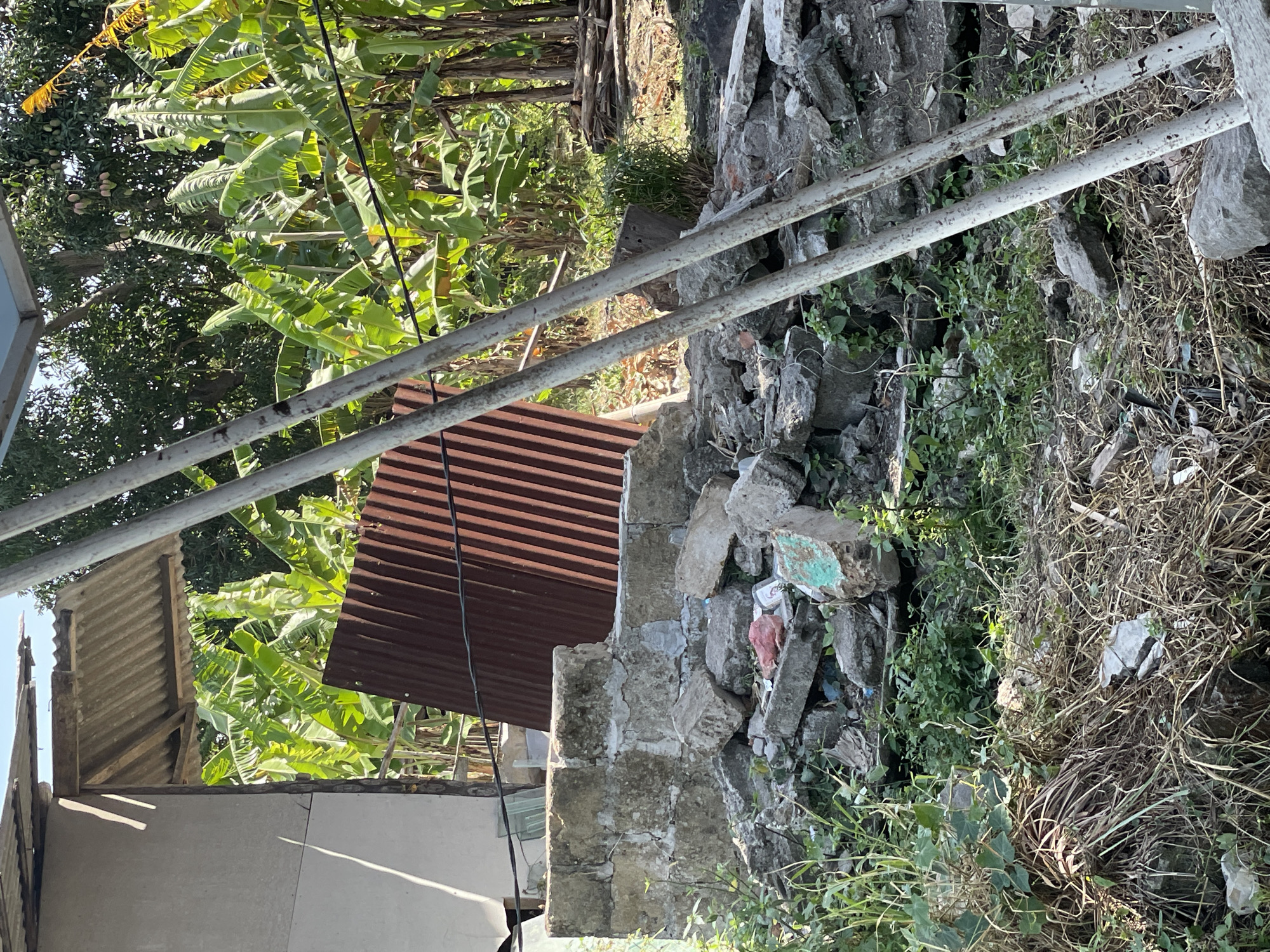 The image depicts a pile of rubble and construction debris in what appears to be a semi-rural or developing area. There is a partial structure on the left with a corrugated metal roof. Behind the rubble, a dense grove of banana trees is visible, suggesting a tropical climate. A rusty corrugated metal sheet leans against the rubble. Two white metal poles, possibly part of a scaffolding or support structure, are prominent in the foreground, angled across the scene. The ground is covered with dry grass and more debris. The lighting indicates it is daytime, likely sunny, with shadows cast by the structures and poles. The location is identified as North Kuta, Indonesia.
