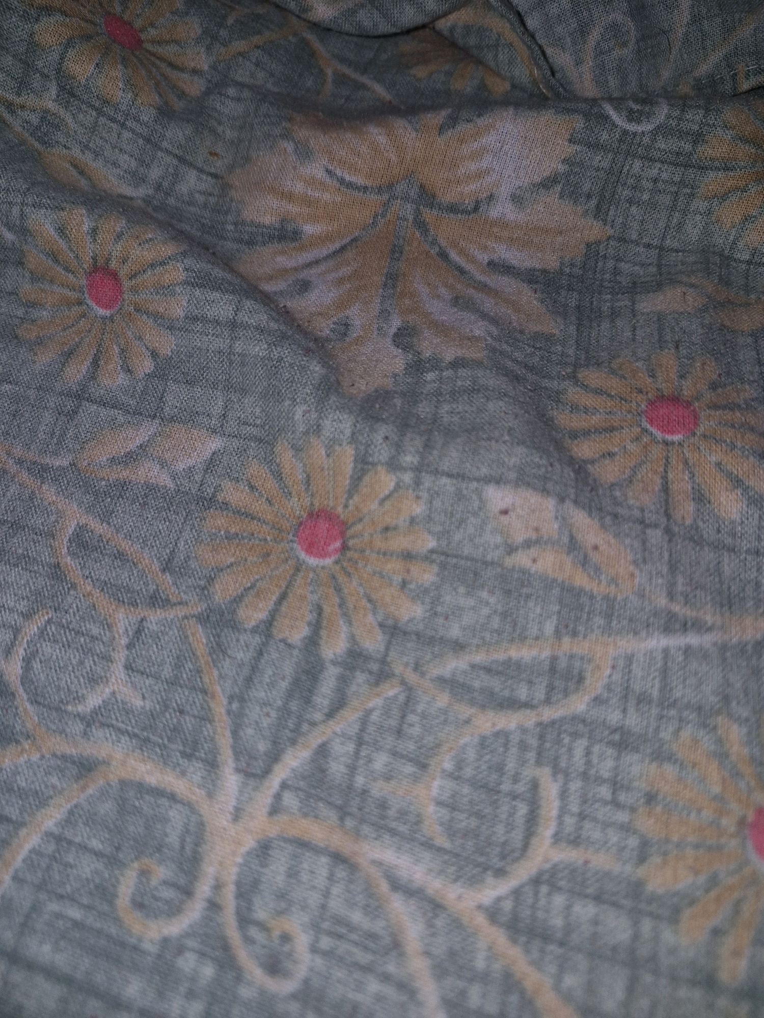 The image shows a close-up of a fabric with a floral pattern. The fabric is draped and folded, creating shadows and highlights that obscure some of the details. The pattern consists of stylized flowers with orange-yellow petals and pink centers, interspersed with leafy motifs and swirling tendrils in a similar color palette. The background fabric is a muted greenish-gray with a subtle texture that appears to be woven. There are no people, animals, or specific landmarks visible. The image is taken from a high angle, looking down at the fabric. This image is not specific to Udalguri, India, as it depicts a generic textile pattern.
