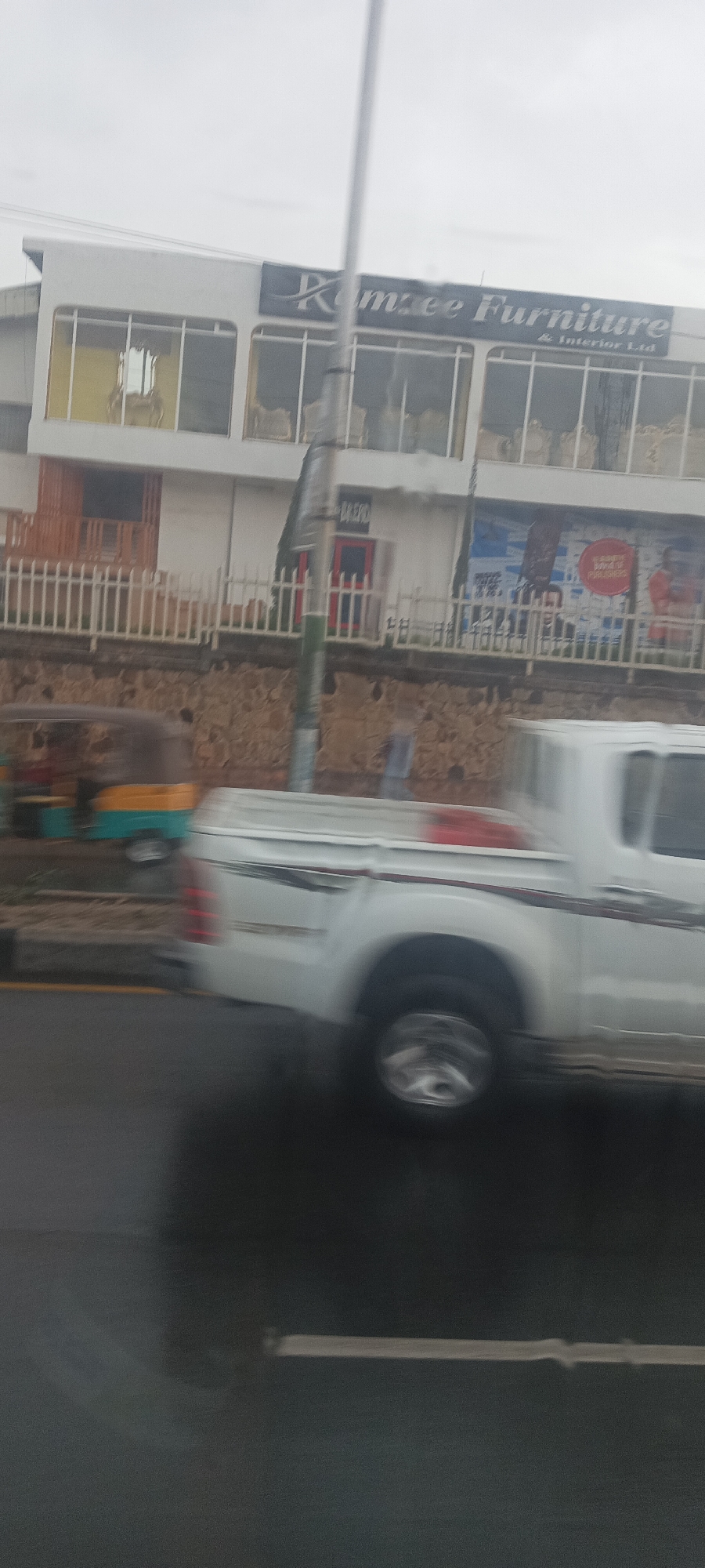 The photo, taken on a Jos, Nigeria street, shows a white two-door pickup truck speeding along a street. A "Komee Furniture & Interior Ltd" building sits in the background along with a three-wheeled tuk-tuk. The scene appears to be from a moving vehicle.
