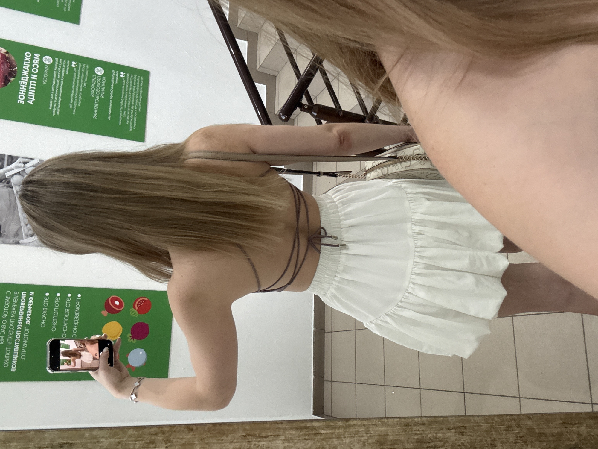 An outdoor shot shows a woman standing in front of a wall covered with posters. The woman is wearing a white skirt and a purple top. She is holding a smartphone in her right hand, and it is pointed towards her reflection in a mirror. The smartphone screen shows a picture of the woman. The wall posters are in Russian. One poster has a picture of a hamburger and french fries. The woman is facing away from the camera, and her hair is down. The background shows a staircase.