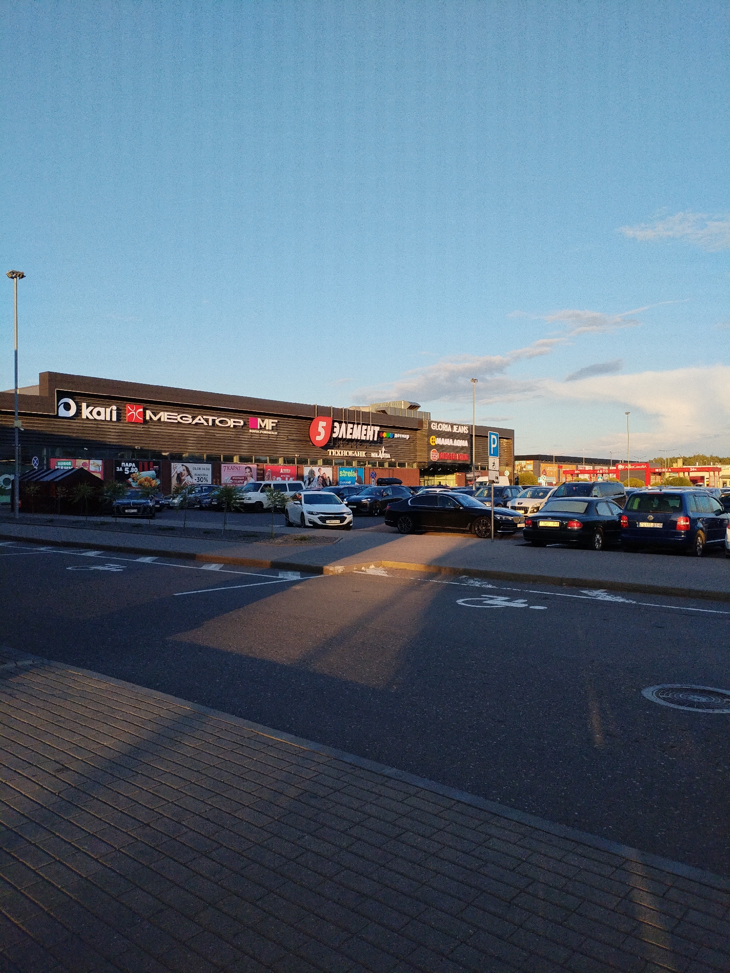 The image captures a commercial complex in Mahilyow, Belarus, under a clear sky with sparse clouds. The complex features a prominent facade with multiple signs, including "kari," "MEGATOP," "MF," and "5 ЭЛЕМЕНТ." Several vehicles are parked in front of the building, suggesting a retail or shopping area. The foreground shows a paved area, possibly a parking lot or walkway, with a distinct pattern of interlocking bricks. The scene appears to be set during the late afternoon or early evening, as indicated by the long shadows cast across the ground. There are no visible people in the image.