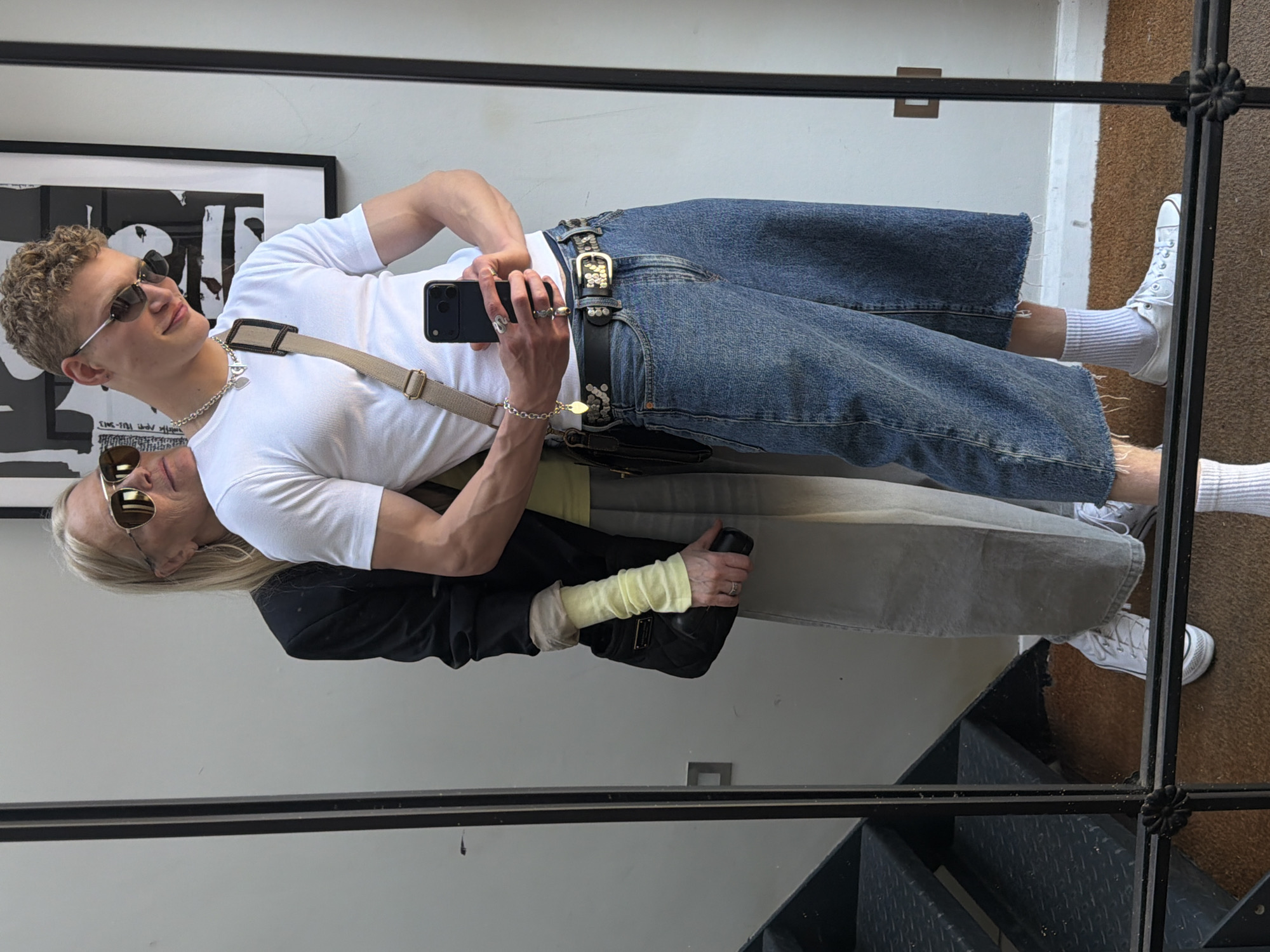 Two individuals, a young man and an older woman, are captured in a mirror selfie taken indoors, likely in London, UK. The image appears to be rotated, showing them horizontally.
The young man, with curly blond hair and sunglasses, is positioned in the foreground. He wears a white ribbed t-shirt, wide-leg blue jeans with distressed hems, and white sneakers with white athletic socks. A black studded belt with a prominent buckle cinches his jeans. He accessorizes with multiple rings on his fingers, a silver chain necklace with a small pendant, and a silver bracelet. A light brown shoulder bag with a wide strap is slung across his chest. He holds a black smartphone, seemingly an iPhone, in his right hand to take the mirror selfie, with his arm loosely around the woman. He has a slight smile.
Behind and partially embraced by the man, the older woman, with long blonde hair and sunglasses, is also seen in the reflection. She wears a white t-shirt underneath a yellow long-sleeved top, layered with a black jacket or blazer. Her lower half is clad in light gray wide-leg pants and white sneakers, also paired with white athletic socks. She carries a black quilted handbag.
The background of the reflection reveals a white wall with a framed piece of art featuring abstract black and white shapes. In the immediate foreground and further back, black metal railings, possibly part of a staircase or landing, are visible. A brown mat or rug is on the floor. The setting suggests a casual, well-lit indoor environment during daytime. No specific text is clearly legible in the image, other than the abstract shapes on the wall art.