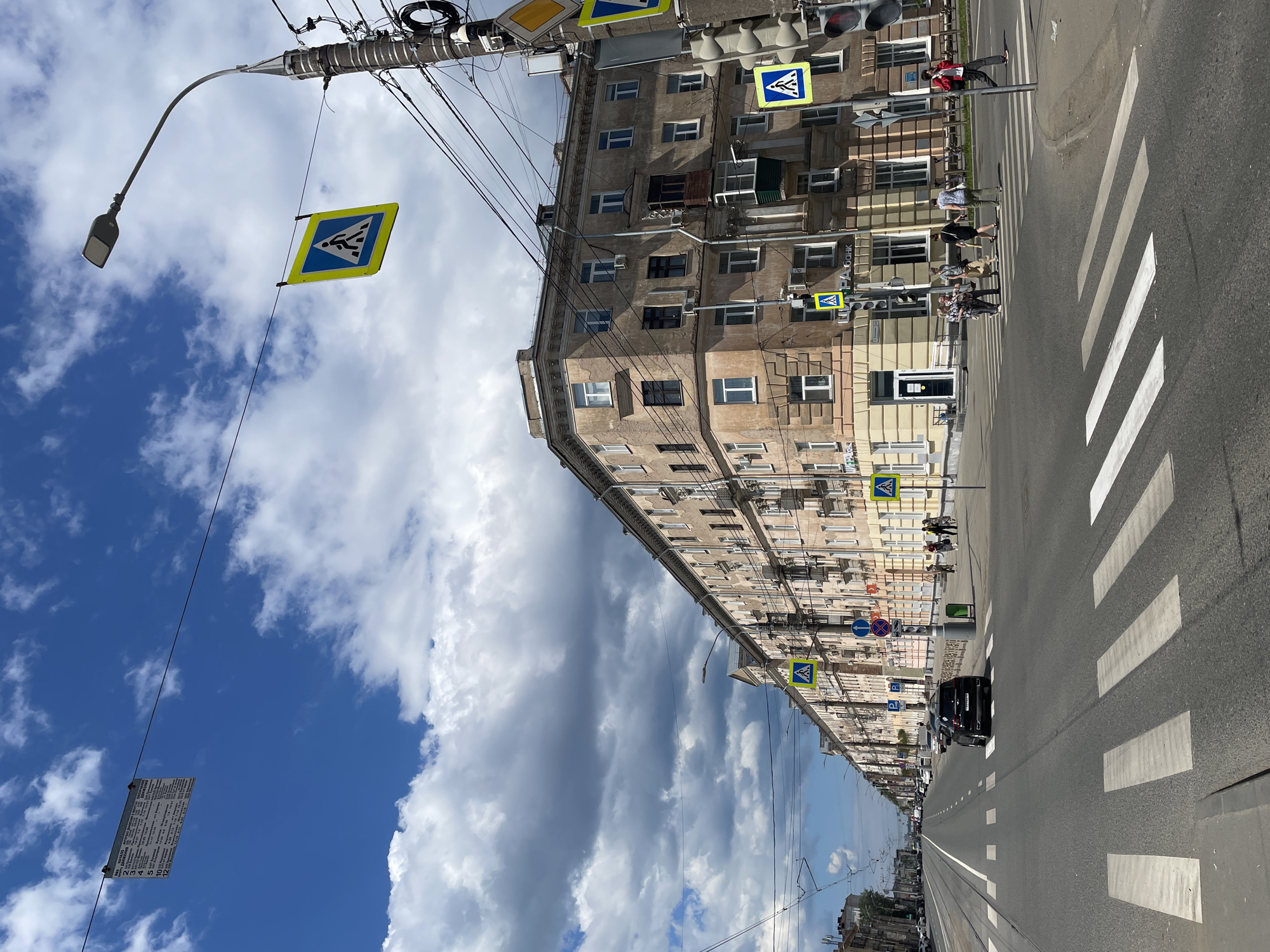 A wide, paved street with tram tracks runs through an urban environment under a bright, cloudy sky. A long, multi-story building with beige and cream-colored facades lines the right side of the street, featuring numerous windows and balconies. Pedestrian crossing signs, yellow and blue, are visible, some hanging from wires and others attached to poles. A black SUV is driving on the street. Several pedestrians are crossing the street at a marked crosswalk in the foreground. Another pedestrian, wearing a red jacket, is walking on the sidewalk on the right. The street appears to be in a state of urban development, with electrical wires and poles visible throughout the scene.