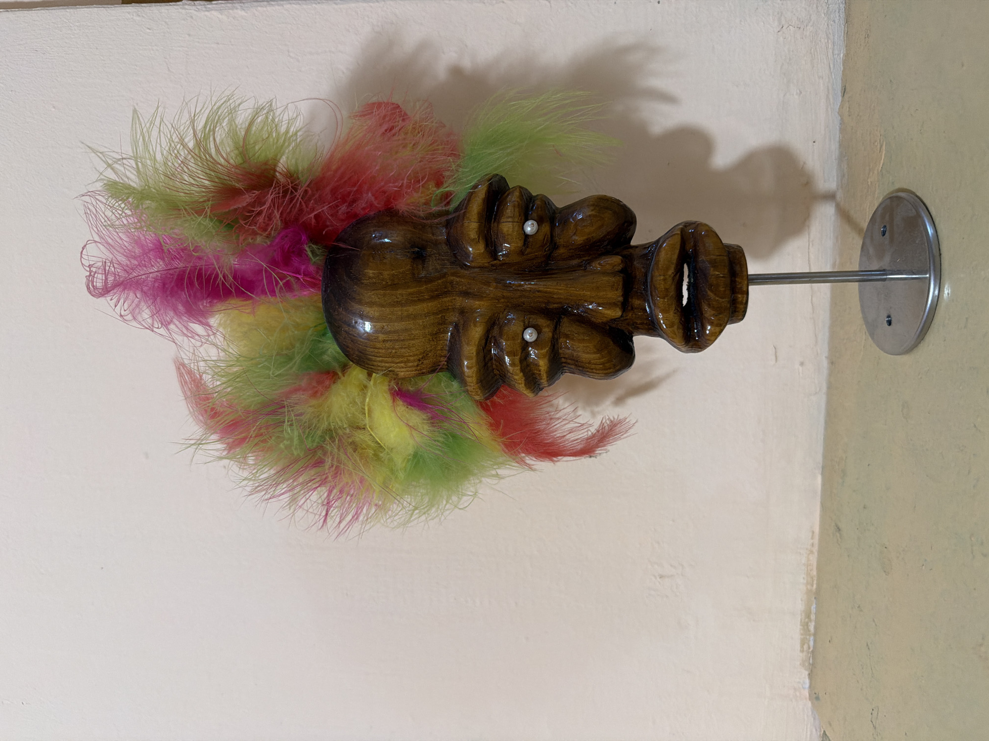 A wooden mask with bright colorful feathers on its head is displayed on a silver stand with a circular base. The mask is brown and has carved features including eyes with small white beads, a nose, and a mouth with an open, carved interior. The feathers are a mix of bright pink, yellow, and lime green, creating a vibrant crest. The mask is positioned in front of a light-colored wall and a tan surface. The scene appears to be indoors, in an area that is not specified beyond being in the city of Unknown, Unknown. There are no people visible in the image, and no actions or interactions are taking place. The lighting casts a shadow of the mask and stand on the wall.