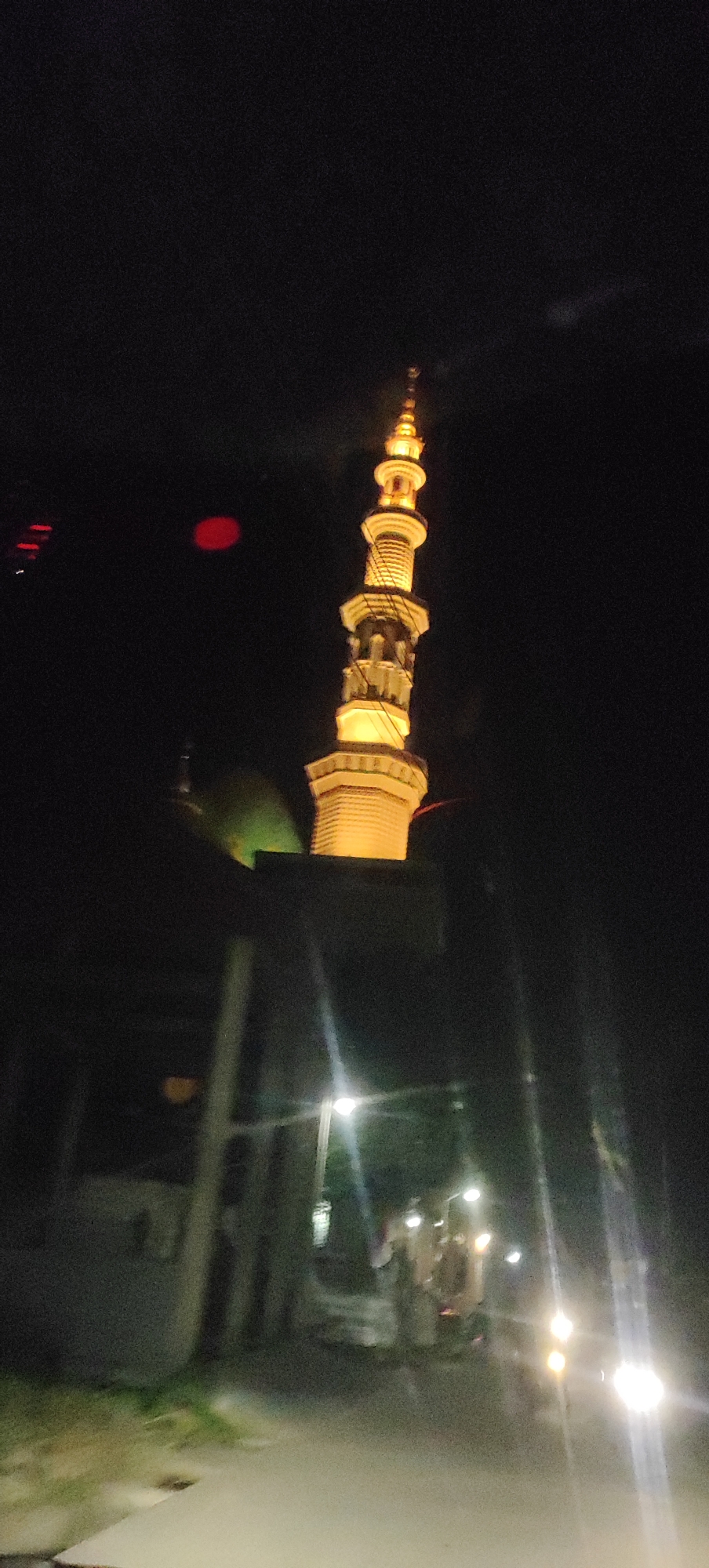 A night-time photograph shows the illuminated minaret of a mosque in Manyar, Indonesia. The minaret, tall and slender, is lit with a warm, yellow glow, accentuating its multiple levels and intricate architectural details. The dark night sky serves as a stark backdrop, with only a few diffused lights visible in the distance. To the left of the minaret, a green-lit dome of the mosque is partially visible. In the foreground, a dimly lit street is depicted, with streetlights casting a soft illumination on the surroundings. The image captures a serene and spiritual ambiance, characteristic of a religious site at night.
