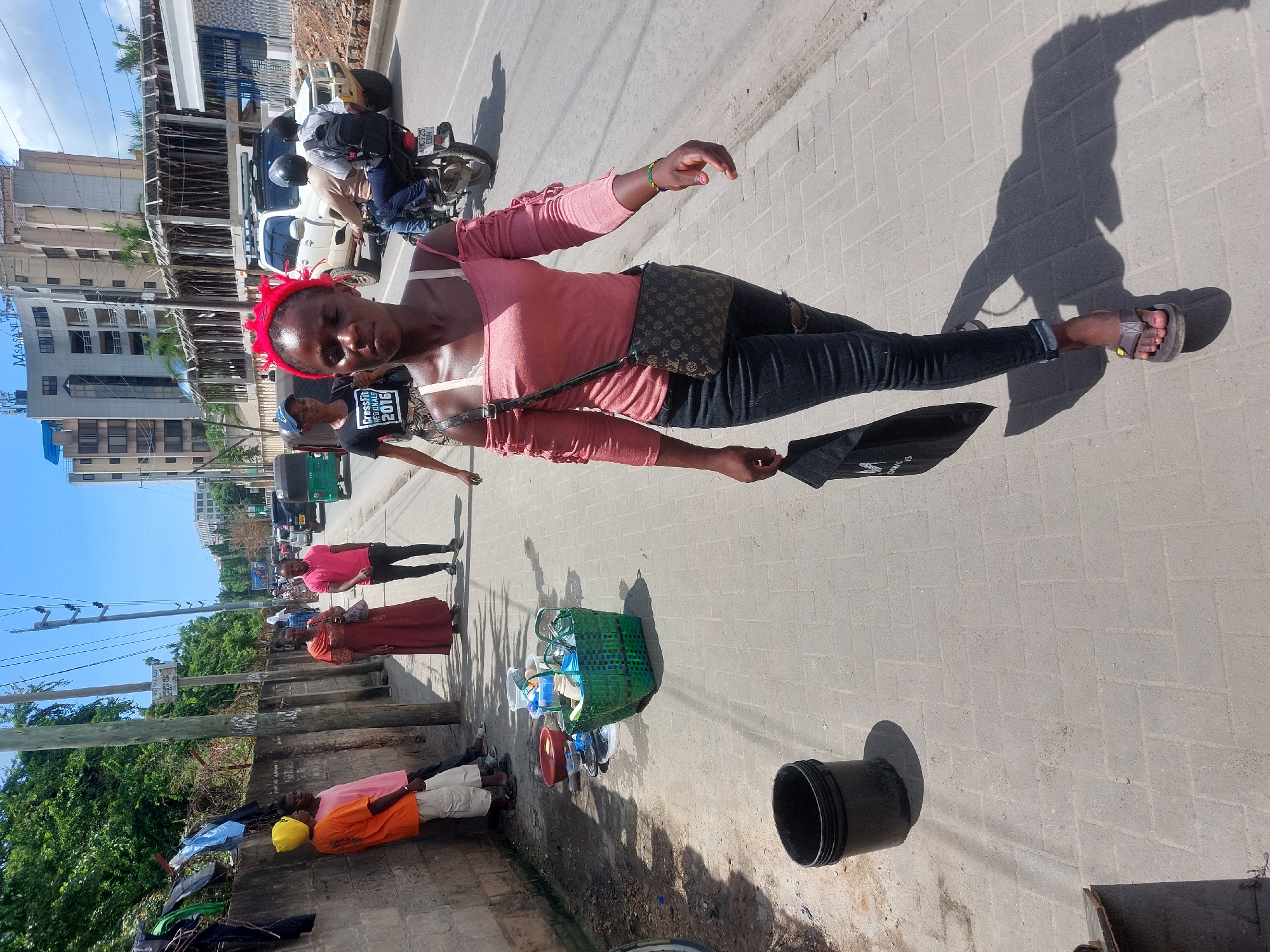 The image captures a lively urban street scene in Dar es-Salaam, Tanzania, under bright, sunny daylight, indicated by long shadows cast on the paved sidewalk. The primary subject is a woman standing towards the right side of the frame, facing slightly towards the viewer. She wears a salmon pink long-sleeved top, dark ripped jeans, and brown sandals with pink nail polish. A red headscarf or bandana is tied around her head, and she carries a small, patterned crossbody bag. In her left hand, she holds a folded black umbrella or similar object.
Behind her, another person is visible, wearing a black t-shirt with the text "CrossFit REGIONALS 2016". Further in the background, the street features vehicles, including a white SUV and a motorcycle with two passengers, one wearing a helmet. The motorcycle's license plate, "MD 923 BBT", is partially visible. Tall multi-story buildings line the street, with the words "MASANAKA BUILDINGS" faintly legible on one structure. Power lines, utility poles, and green tropical foliage are also present.
Along the sidewalk, several other people are seen, some standing or walking, suggesting a busy street environment. A green woven basket filled with various plastic household items such as containers and cups, alongside a red basin, is placed on the ground, indicating street vending. A black bucket is also situated nearby. The scene depicts everyday life and commerce on a bustling city street.