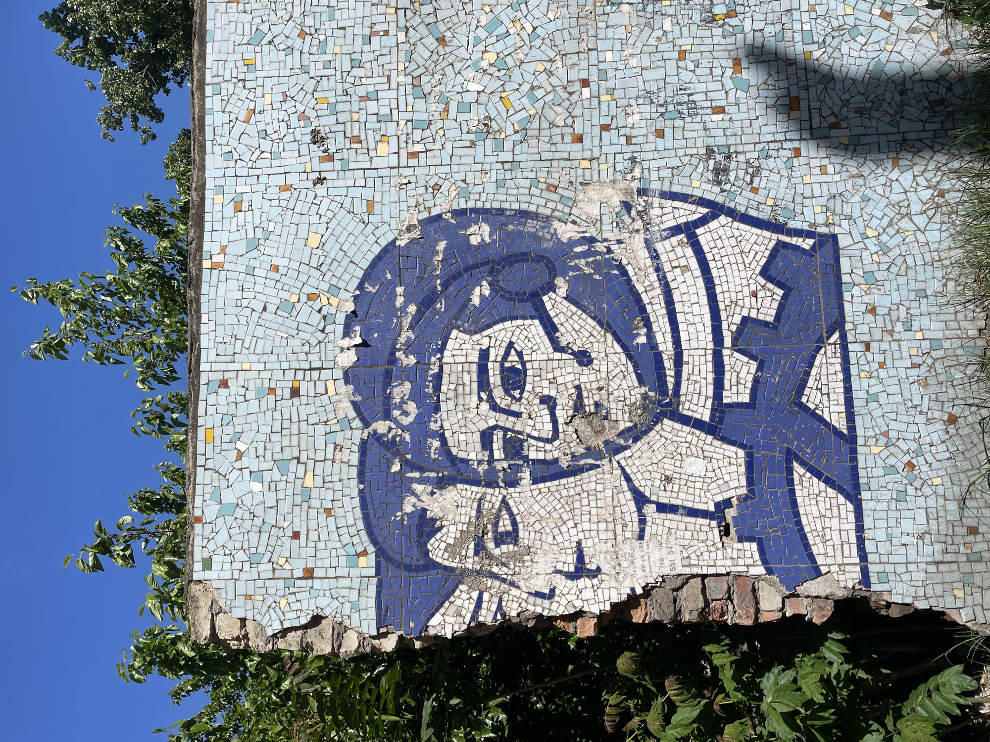 The image depicts a decaying mosaic mural on a building in Tbilisi, Georgia. The mural, constructed from small square tiles, features a stylized illustration of a person wearing a helmet or head covering, rendered in blue and white. The surrounding background mosaic consists of light blue, yellow, and brown tiles. The bottom edge of the mural is broken and crumbling, revealing a brick foundation. Vegetation, including trees and bushes, partially obscures the mural and its base. The sky is visible in the background, transitioning from blue to a lighter hue.