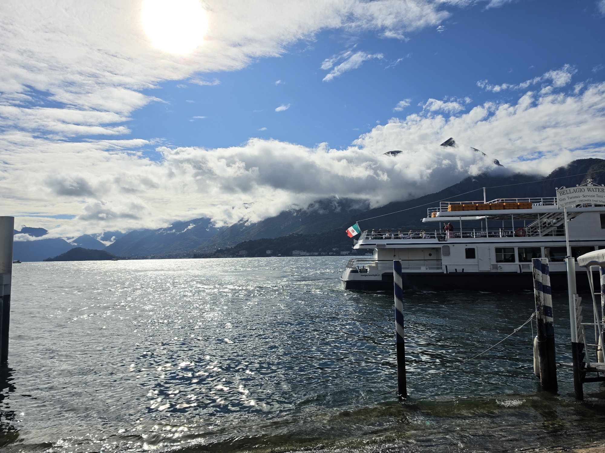 The image depicts a sunny day on a body of water, likely a lake, with a passenger boat docked at a pier. The water is choppy and reflects the sunlight, creating a shimmering effect. In the background, lush green mountains rise, partially obscured by soft white clouds that are catching the sunlight. The sky is a vibrant blue, with the sun positioned in the upper left quadrant, casting a bright glare.
A white passenger boat is moored to a pier in the foreground. The boat has multiple decks, with windows and life vests visible. An Italian flag flies from the upper deck. Several people are visible on the boat's decks, some standing and some sitting. The pier is marked by striped bollards in the water, and a section of a walkway or structure is visible on the right. A sign on the boat reads "BELLAGIO WATER TAXI" and provides contact information. The foreground of the image shows the edge of the land with some rocks and shallow water.
The scene appears to be a scenic waterfront location in Italy, likely a popular tourist destination. While the image doesn't explicitly state the location is San Giovanni, Italy, Bellagio is a well-known town on Lake Como in Lombardy, Italy, and this scenery is consistent with that region.