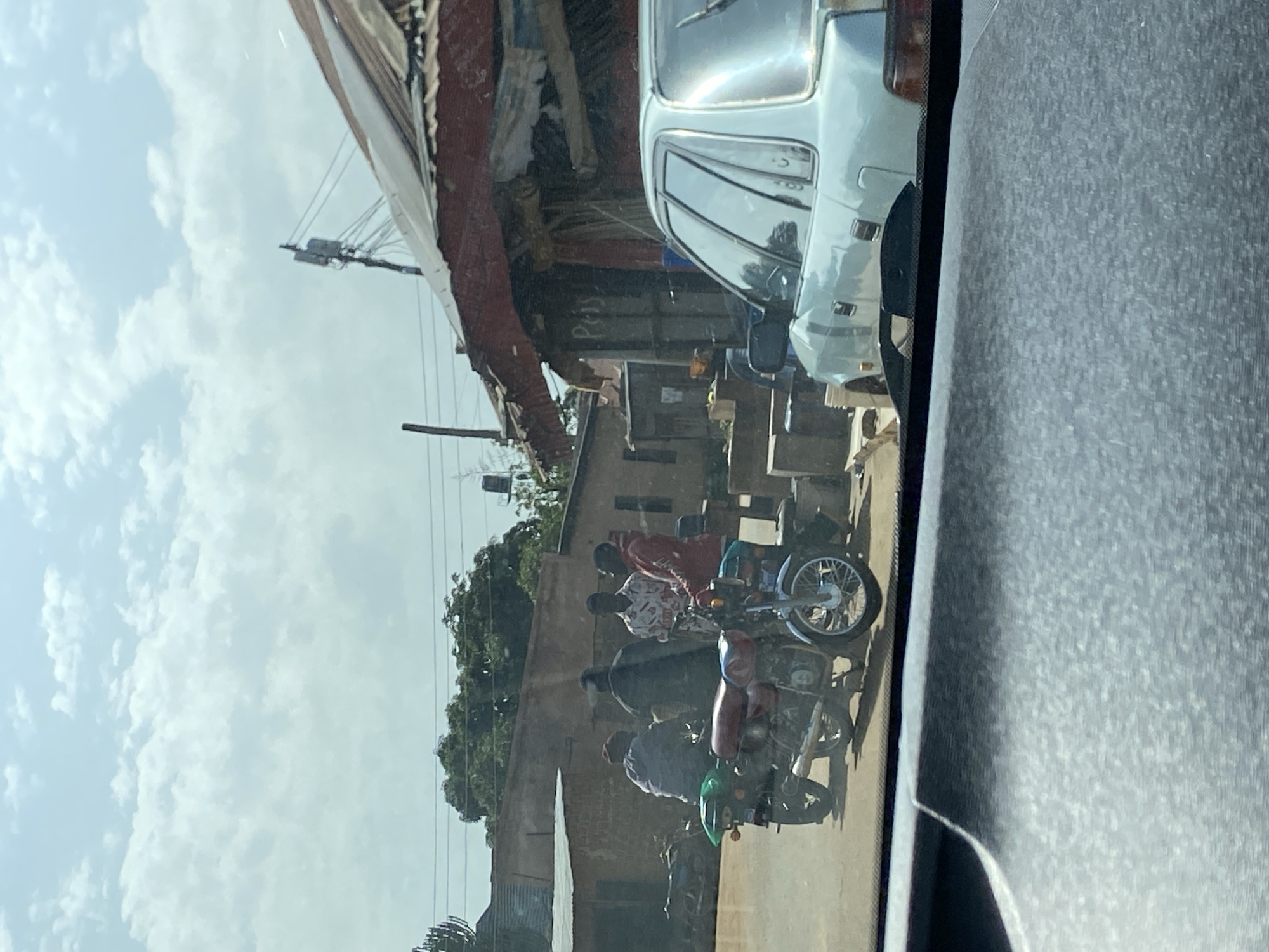In Bar Jirgi Summa, Nigeria, four individuals are gathered around two motorcycles on a dusty road. Two motorcycles are parked side-by-side in the foreground, one a red and black model with a green rear fender, and the other a blue and silver model. Three men are standing and interacting near the motorcycles, while a fourth person sits on the red and black motorcycle.
Behind the motorcycles, a pale blue sedan is parked. In the background, there are several low-lying buildings with corrugated metal roofs and brick or mud walls, suggesting a mix of residential and commercial structures. A telephone pole with wires extends into the sky above the buildings. Trees with dense foliage are visible in the middle ground. The sky is partly cloudy with scattered white clouds against a light blue backdrop. The scene appears to be a typical street view in a Nigerian town.