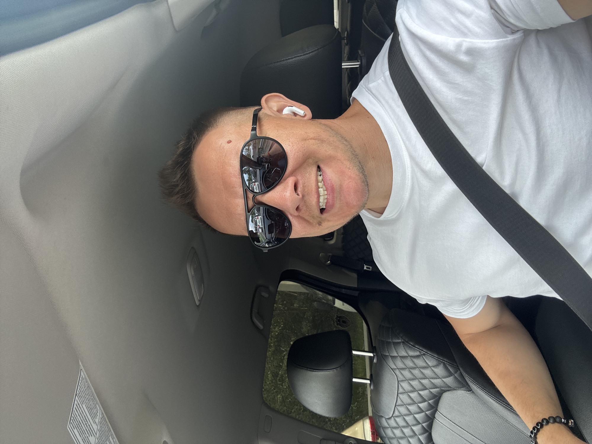 The image shows a man in a white t-shirt and sunglasses, sitting in the driver's seat of a car. He is smiling and looking at the camera. He is wearing white wireless earbuds. The interior of the car appears to be black leather with a quilted pattern on the seats. A seatbelt is visible across his chest. The background is out of focus, but suggests an outdoor setting with greenery. The lighting suggests it is daytime, possibly sunny. The image appears to be a selfie taken by the man. There is no visible text in the image.
