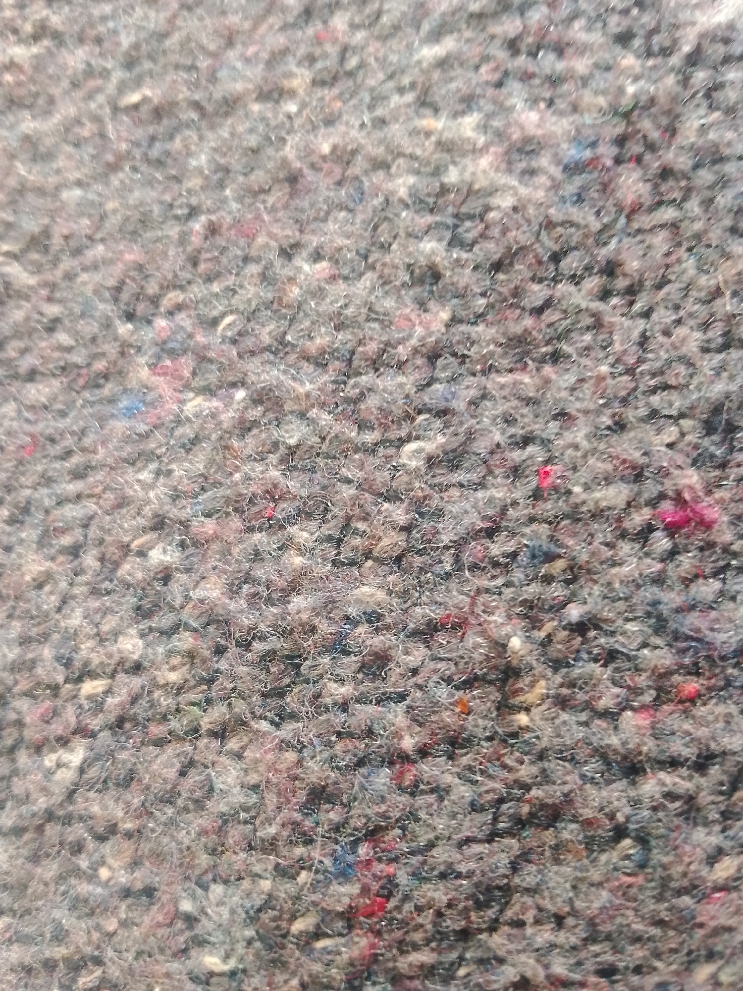 The image is a close-up, overhead view of a textured, multi-colored textile. The dominant color is gray, interspersed with flecks of red, pink, blue, and possibly white fibers. The texture appears to be made of coarse, tangled threads or fibers, giving it a rough and somewhat uneven surface. Some areas show more prominent clumps of color, particularly bright red and pink. The lighting suggests it is outdoors, with sunlight casting subtle shadows that highlight the texture. There are no discernible objects, people, or specific environmental features visible that would indicate a location within the city of Jalingo, Nigeria, or any other specific location. The image solely captures the detail of the textile itself.