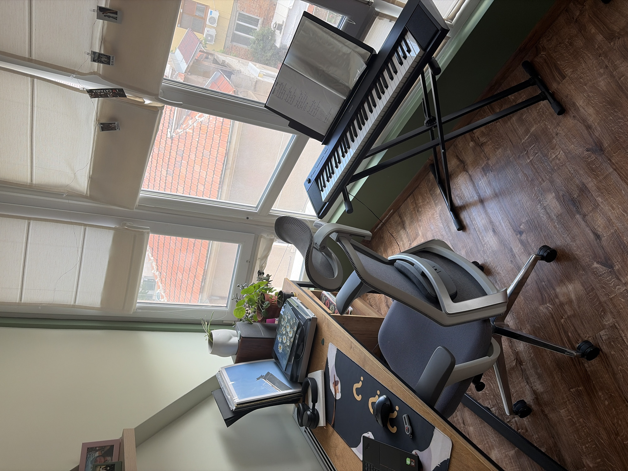 The image depicts a home office or music studio setup against a window. A wooden desk is positioned to the left, featuring a laptop, a computer mouse, a USB drive, headphones, a turntable with records, and a stack of vinyl records. A grey office chair with wheels is placed in front of the desk. To the right of the desk, a black keyboard is set up on a foldable stand, with sheet music displayed on a music stand. The background shows a window with white blinds, revealing buildings and a tiled roof outside. The room has light green walls and a wooden floor.