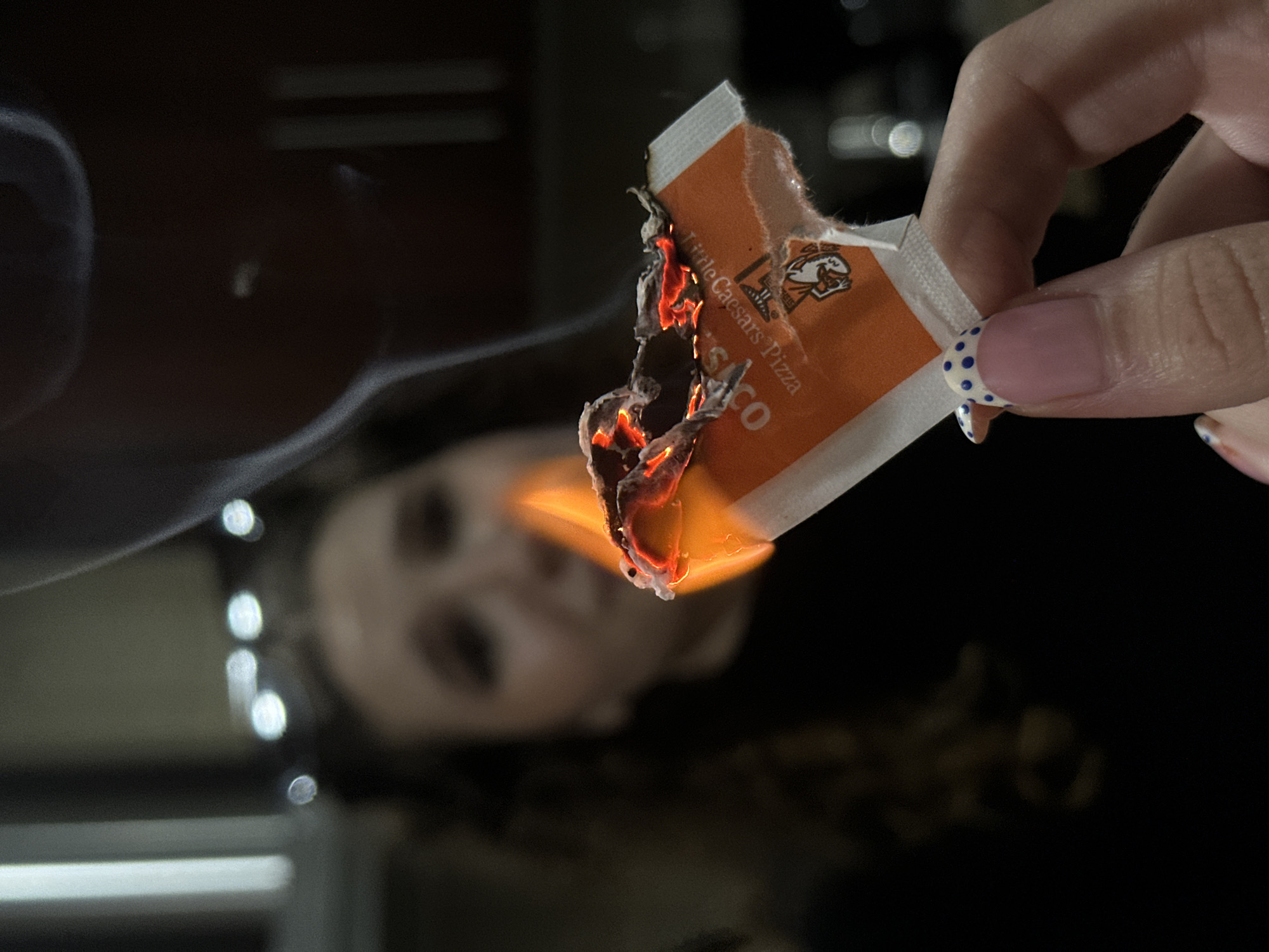 A person's hand holds a small, burning piece of paper with an orange logo and text that reads "Little Caesars Pizza." The flame is bright orange and has glowing embers. Smoke is rising from the burning paper. In the background, a blurred figure of a person is visible. The environment appears to be indoors, with dark cabinetry and lights visible in the background. This scene takes place in San Bernardino Tlaxcalancingo, Mexico.