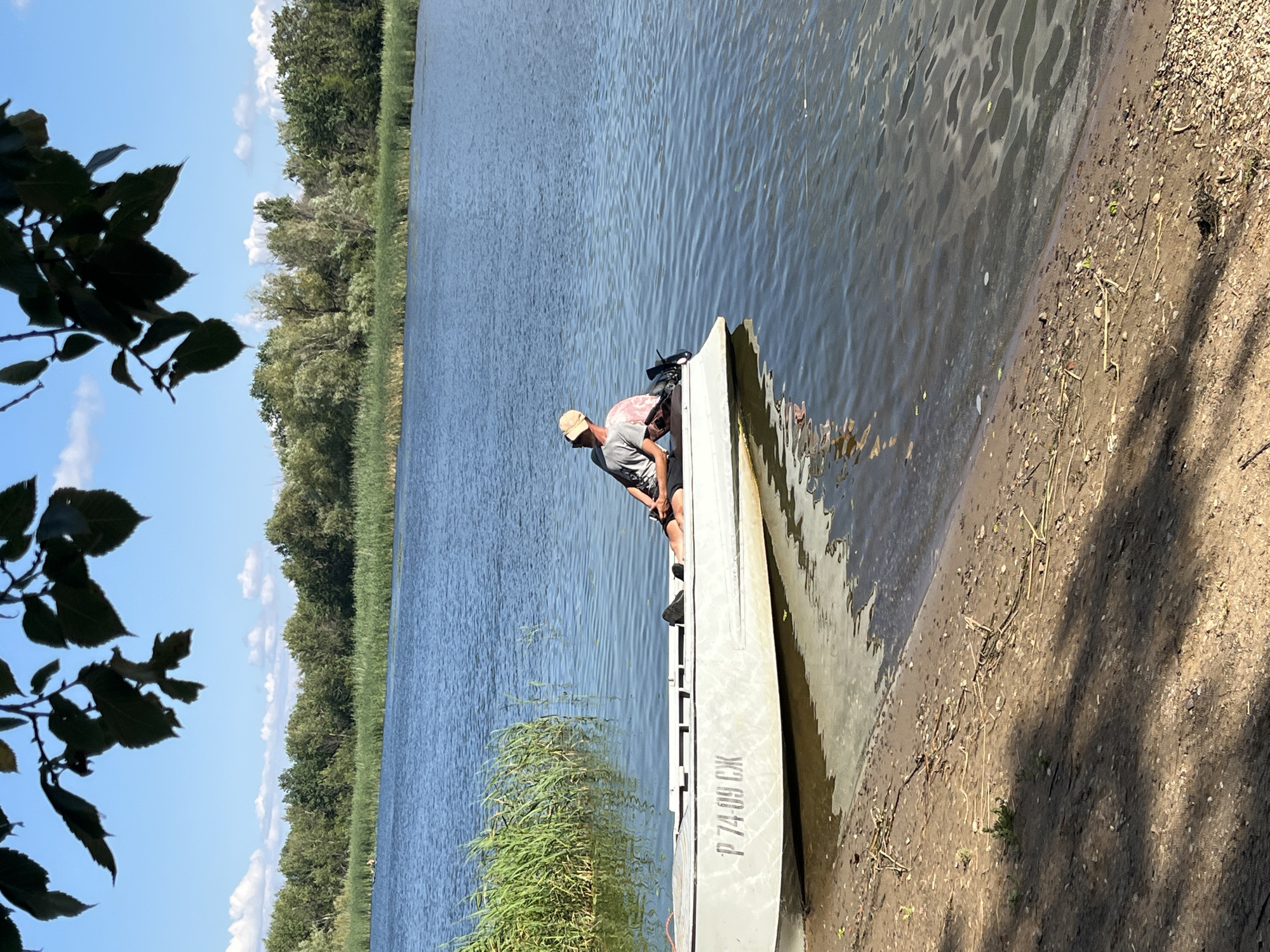 A person wearing a light-colored baseball cap and a grey t-shirt sits in a light-colored boat docked on a sandy shore. The boat is positioned at an angle to the shore, with its bow pointing towards the water. The person is facing away from the camera, looking out at a body of water. Their legs are extended, and they are holding a fishing rod. The boat has Cyrillic lettering visible on its side, "P 74-09 GX". The background consists of a dense line of trees and vegetation along the opposite bank of the water. The sky is blue with scattered white clouds. The water is calm with ripples. The sandy shore is littered with small stones and dry plant debris. The overall scene suggests a peaceful, outdoor setting, possibly a river or lake. This image was taken in Saratov, Russia.
