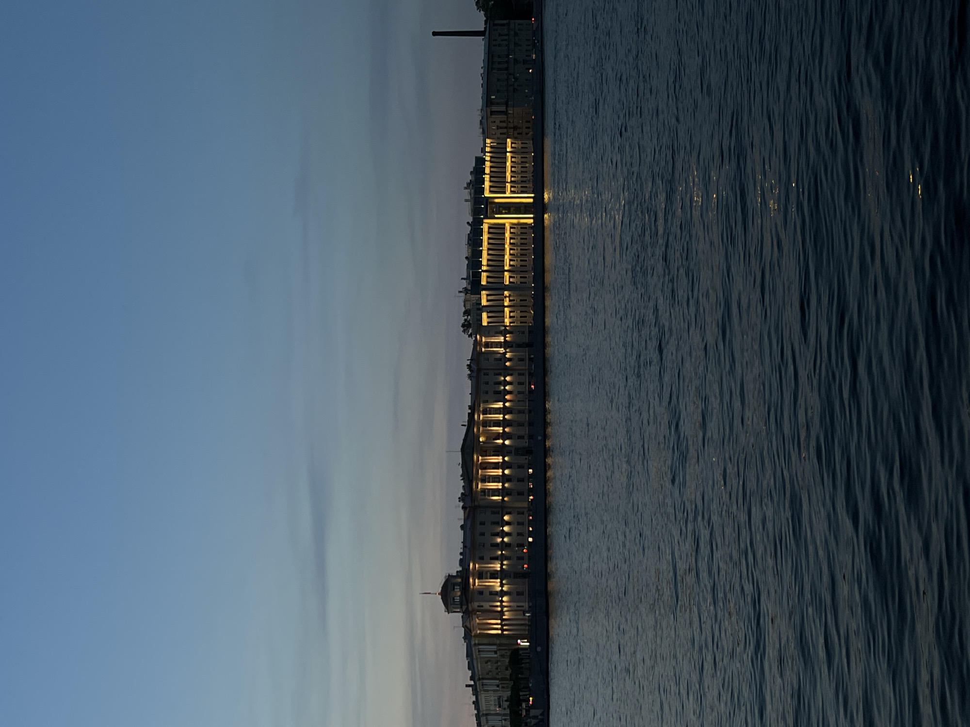 An outdoor, vertical, eye-level shot captures a long, illuminated building situated along a body of water at dusk. The building is neoclassical in style, with a symmetrical facade featuring columns and arched windows. Numerous lights illuminate the facade, casting a warm glow. A dome structure with a flagpole sits atop the left section of the building, and a tall, dark smokestack is visible on the right. The body of water in the foreground is dark and choppy, reflecting the lights of the building. The sky above is a gradient of deep blue and purple, with subtle cloud formations. The overall scene depicts a historic urban architectural landscape at twilight.
