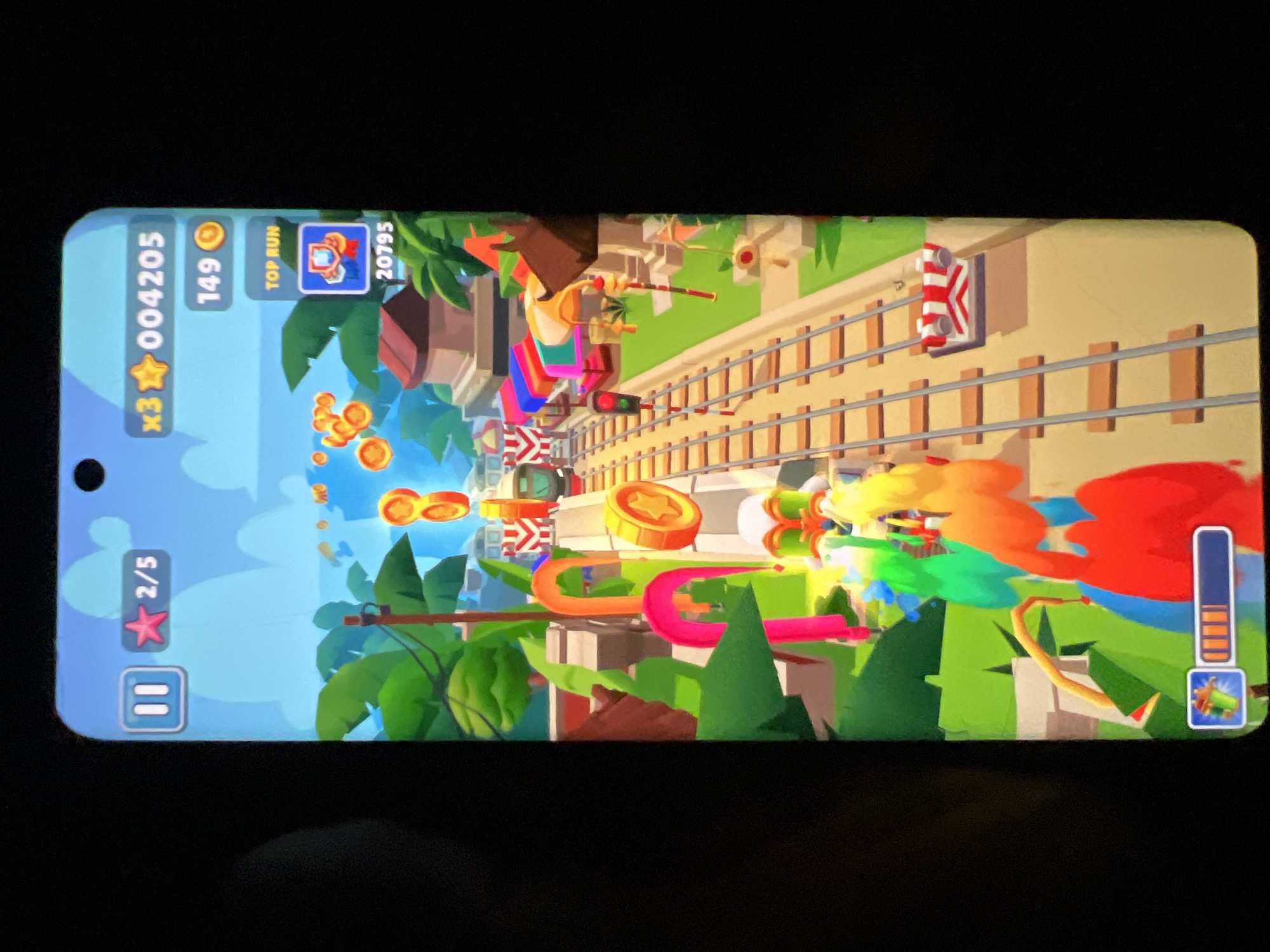The image displays a screenshot of the mobile game Subway Surfers. The game is set in a vibrant, tropical environment with lush green foliage and traditional-looking buildings. The player character, a colorful, abstract figure, is depicted running along train tracks, collecting coins and avoiding obstacles. In the top left corner, a pause button and a star icon with "2/5" indicate progress in a game challenge. To the right, the player's score is shown as "x3 004205" with "149" coins. Below this, "TOP RUN" with a trophy icon and "20795" suggests a leaderboard or high score feature. The game's visual style is cartoonish and energetic, typical of mobile games. The setting appears to be a stylized representation of a city, but no specific landmarks or elements clearly identify it as Lagos, Nigeria. The overall scene is one of active gameplay within a colorful, animated digital world.