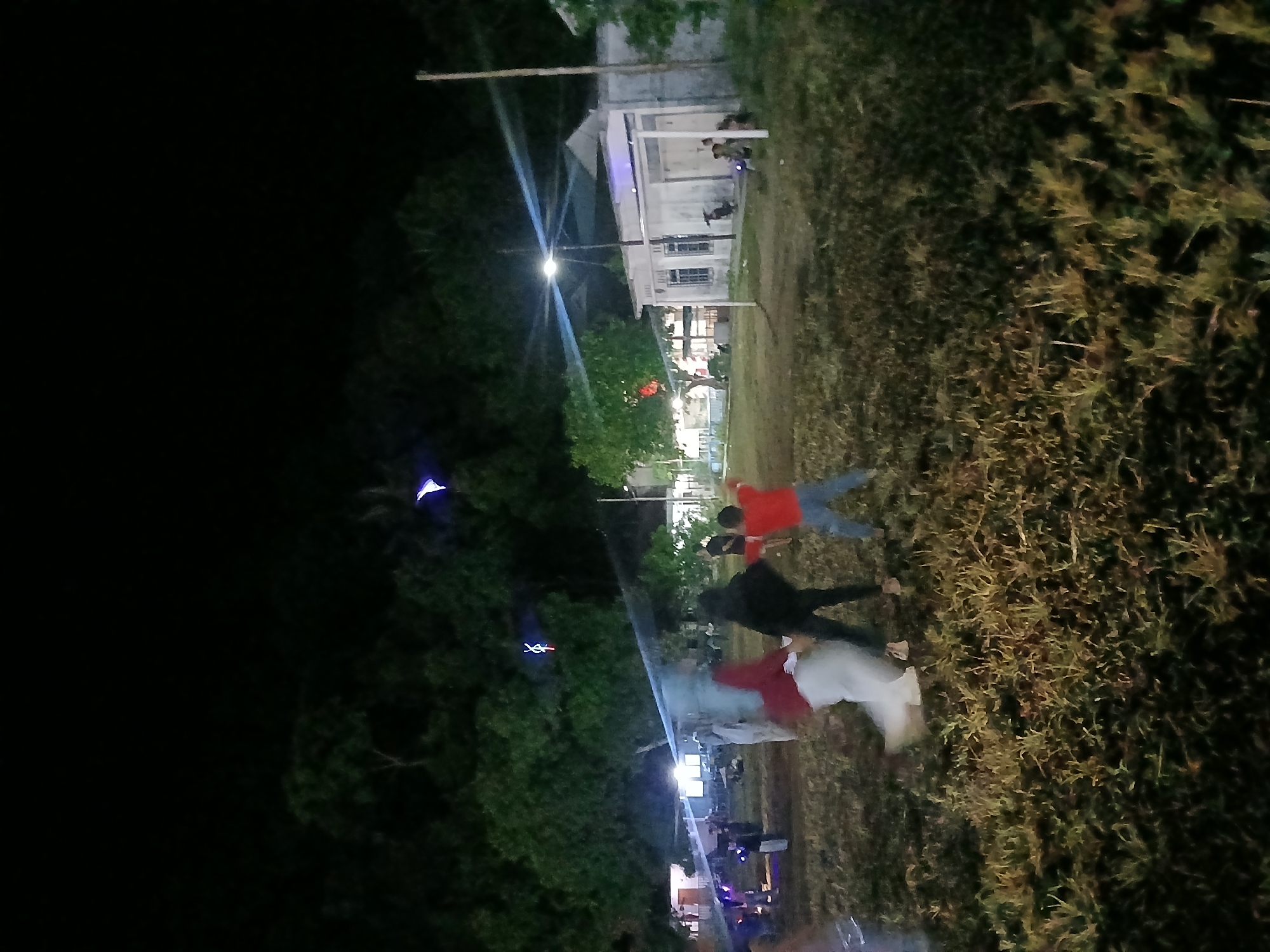 The image depicts a nighttime outdoor scene, likely in Terempa, Indonesia, with a group of people gathered in a grassy field. Several individuals are in motion, suggesting activity or movement. In the foreground, the ground is covered in dry grass and some scattered foliage. In the midground, a cluster of people are visible. One individual appears to be wearing a red shirt and blue pants, with arms outstretched. Another figure is in a black shirt and dark pants, with their back to the camera. A third person, partially obscured and appearing to be in motion, is wearing a red head covering and white garment. Further back, more figures are present, some standing and others seemingly engaged in conversation or observation.
The background features several structures, including what appears to be a building with a white facade and a covered porch, possibly a house or community building. Lights are visible from within and around these structures, illuminating the area. Trees with dense foliage line the upper portion of the image, with the dark sky above. Several lights and some colorful illuminated objects, possibly lanterns or decorative lights, are suspended in the air amidst the trees. A tall, slender pole, possibly a utility pole or flagpole, stands to the right of the frame. The overall lighting suggests an event or gathering taking place after dark. The scene conveys a sense of community or festive activity in a semi-rural or suburban environment.