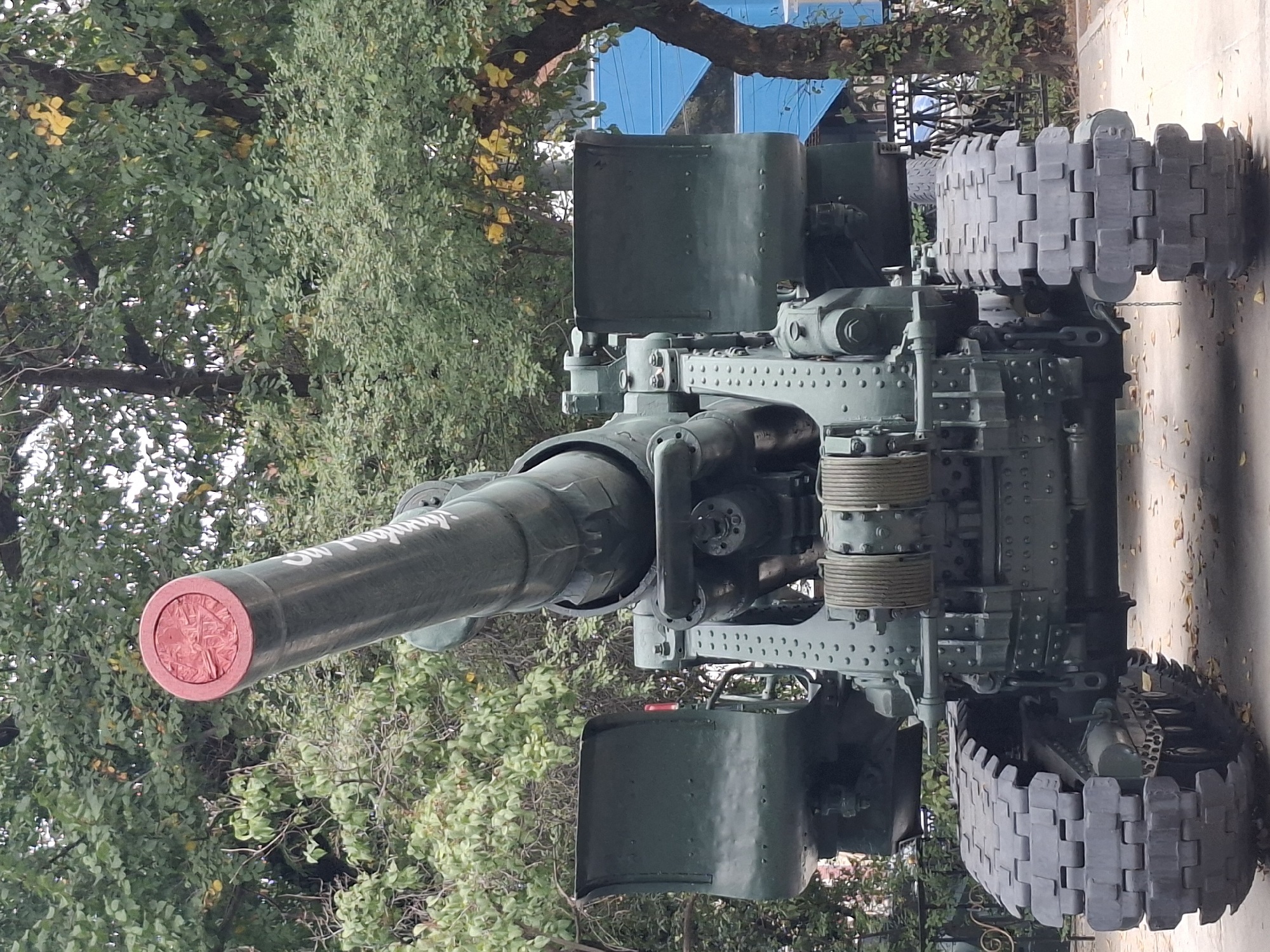 The image shows a full frontal view of a large, olive-drab artillery piece, likely a howitzer, with caterpillar tracks. The barrel of the cannon is elevated and pointed upwards. The end of the barrel is capped with a red circular object, which appears to have some kind of embossed design. White lettering is visible along the side of the barrel. The artillery piece is positioned outdoors, with lush green trees forming the background. Some yellow leaves are visible on the trees, suggesting it might be autumn. In the background, to the right, a bright blue structure is partially visible behind a fence, and further to the right, a portion of a tree trunk with ivy is present. The ground beneath the artillery piece is paved, with some fallen leaves scattered on it. The scene is set in a park-like environment. This artillery piece is on display at Panfilov Park in Almaty, Kazakhstan. There are no people visible in the image.