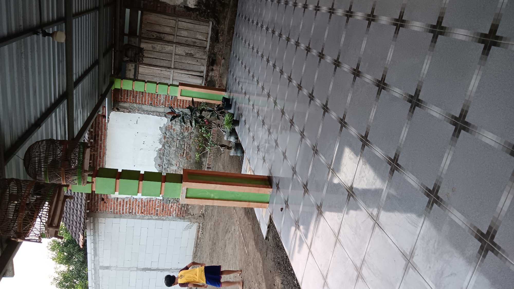 This image shows a scene in Sonobekel, Indonesia, featuring an outdoor tiled patio area leading to a building with a covered roof supported by green and orange pillars. Two ornate birdcages are suspended from the ceiling. A young child, wearing a yellow shirt and blue shorts, is walking away from the camera on a dirt path to the left of the patio. Several potted plants are placed along the edge of the patio. The background shows a brick wall, a white-washed wall, and a wooden door. The overall impression is a residential or semi-residential outdoor space.