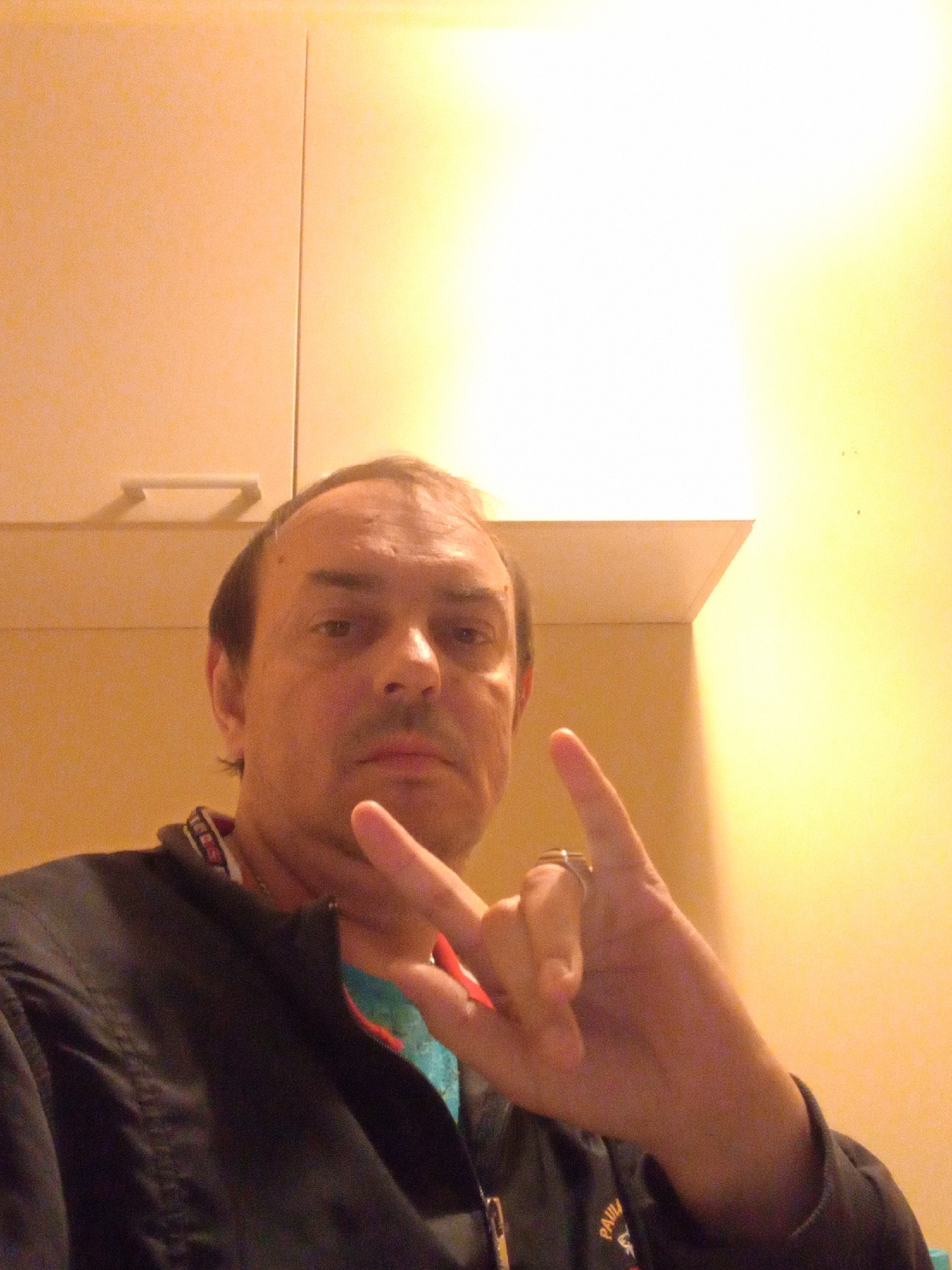A Caucasian male, appearing to be in his 40s, is captured in a selfie-style photograph. He has short, dark hair, a visible hairline, and appears to be clean-shaven or with light stubble. His expression is neutral. He is wearing a black jacket with a collar that has a repeating pattern of text and symbols. Beneath the jacket, a blue garment is partially visible.
The man is positioned in what appears to be an indoor setting. Behind him, a light-colored wall is visible, with a horizontal line suggesting cabinetry or a shelf. To the right, the wall transitions to a bright yellow hue, illuminated by an overhead light source. The lighting casts a distinct shadow and a bright, blown-out area on the wall.
The man's right hand is raised, with his index and pinky fingers extended, forming a 'rock on' gesture. A ring is visible on his right ring finger. His left hand is not clearly visible. The overall image has a slightly grainy quality, possibly due to low light conditions or camera settings.
There are no explicit visual cues in the image that indicate the location is within the city of Kajaani, Finland. The provided environment could be a residence, an office, or another indoor space.