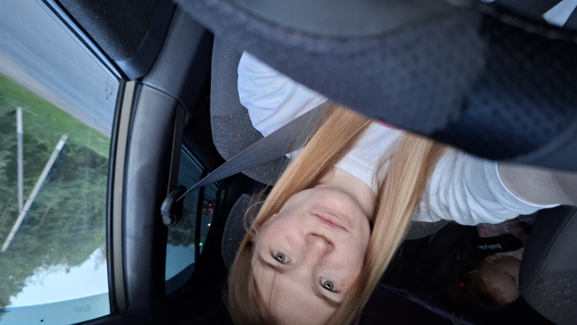 A selfie taken inside a car by a fair-skinned woman with long, light-reddish-blonde hair. The woman is wearing a white t-shirt and a seatbelt. The steering wheel is visible at the top of the frame. Outside the window, there is a view of greenery and a roadside. In the back seat, a baby in a car seat is seen. The location is likely near Chaikovsky, Russia, based on the roadside environment and the angle of the selfie.