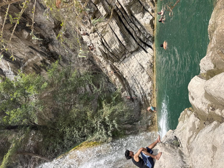 This image depicts a natural swimming area located in San Miguel Atlapulco, Mexico, under bright daylight conditions. A large waterfall cascades from the upper left into a clear, teal-colored natural pool that occupies the lower half of the frame. The left side of the scene features the waterfall, its base surrounded by large, light-colored rocks. Dense green vegetation and trees cling to the rocky cliffs above and to the left of the waterfall.
The right side of the image is dominated by steep, layered rocky cliffs, showing distinct horizontal stratification. Scattered vegetation, including some dry brush, grows on these cliffs.
Approximately ten individuals are present. In the immediate foreground, a young male with dark hair, wearing a black tank top and blue and white shorts, sits barefoot on a large, light-colored rock. He has an ankle bracelet on his left ankle and is looking towards the right side of the pool. His feet are positioned near the water's edge, close to a dark rope that extends across the water towards the right.
Within the pool, several individuals are swimming or wading. One male in light-colored shorts stands in the shallow water near the base of the waterfall, facing away from the camera and creating a splash. Further into the pool, at least four other individuals are visible; one male with a bare back is submerged to his shoulders, and two individuals (one male and one female) are positioned closer to the far bank, appearing to interact. Another person's head is partially visible in the water.
On the steep rocky cliff face to the right, multiple individuals are engaged in climbing or descending. Three males are distinctly visible on the rock layers: one higher up, wearing dark shorts; another below him, also in dark shorts; and a third further down, appearing to use a rope for assistance or support. Another individual is visible near the water's edge at the base of the cliff. The rope seen spanning the water in the foreground also connects to the cliff face on the right, and another rope extends down the cliff face towards the water.