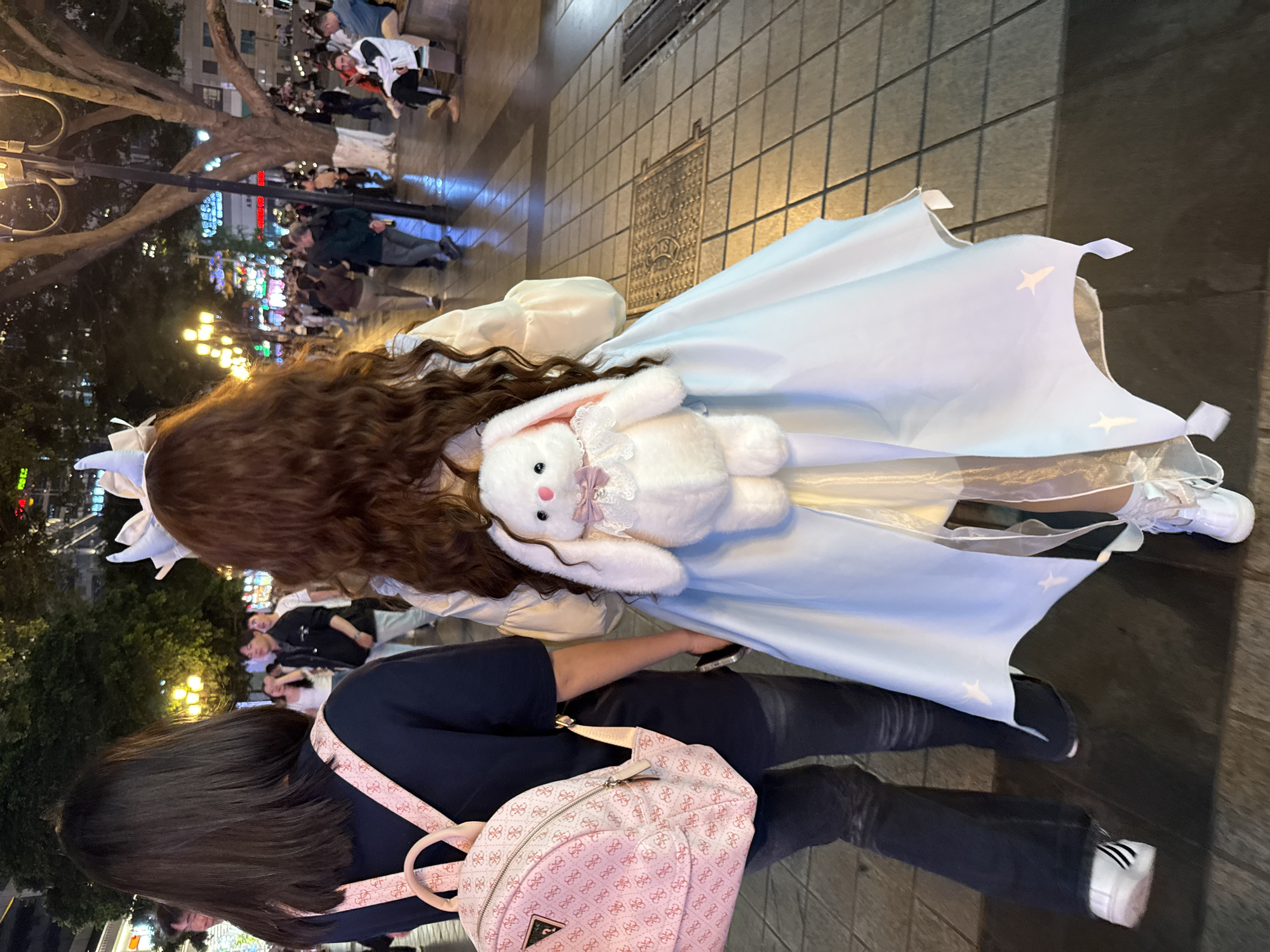 The image, taken from above, shows a person in a light blue dress with a white stuffed rabbit. The dress has star details. Next to them, another person has a pink backpack. They are standing on a paved street that seems to be crowded, with people visible in the background. Street lights are visible, indicating it is nighttime. The location is Yuzhong District, China.