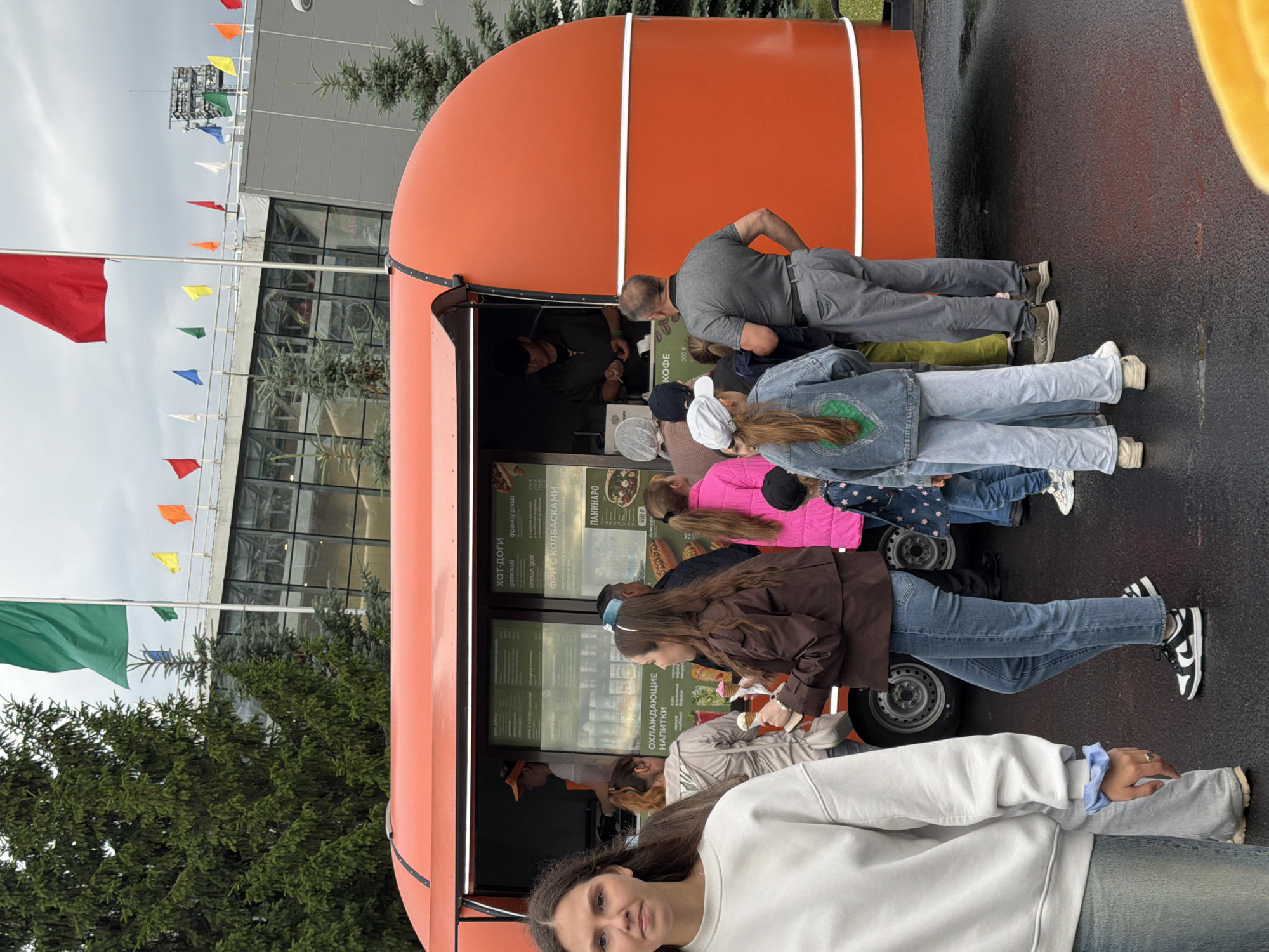 An outdoor scene captures an orange food truck with a menu displayed in its serving window. Several people are in line to order food and drinks. A young woman in a white sweatshirt and light blue jeans stands in the foreground, facing slightly away from the camera. Behind her, others are queuing, some holding ice cream cones. The food truck is parked on a wet, dark surface, possibly asphalt, indicating recent rain. In the background, a large building with a glass facade is visible, along with several small flags of various colors fluttering from poles. Mature coniferous trees are also present in the background. The sky is overcast.