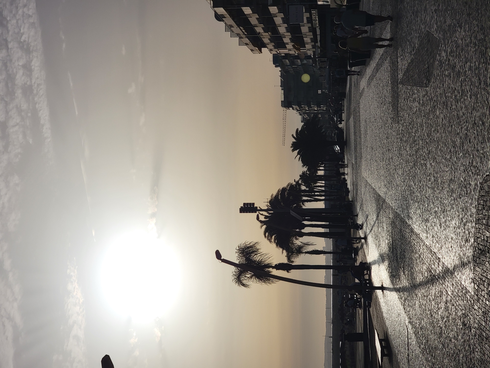 The image depicts a sunlit promenade in Quarteira, Portugal, likely during late afternoon or early evening given the low sun angle and long shadows. The promenade is paved with cobblestones that reflect the bright sunlight. A line of silhouetted palm trees runs along the left side of the promenade, casting long, distinct shadows across the paving stones. In the background, a modern building with multiple balconies dominates the right side of the scene, with a construction crane visible further down the street. Several people are walking along the promenade, mostly in silhouette, including a couple holding hands in the foreground. There are benches and streetlights along the promenade. The sky is hazy with some scattered clouds, and the sun is a bright, diffused orb near the horizon. The overall atmosphere is one of a warm, coastal urban environment.