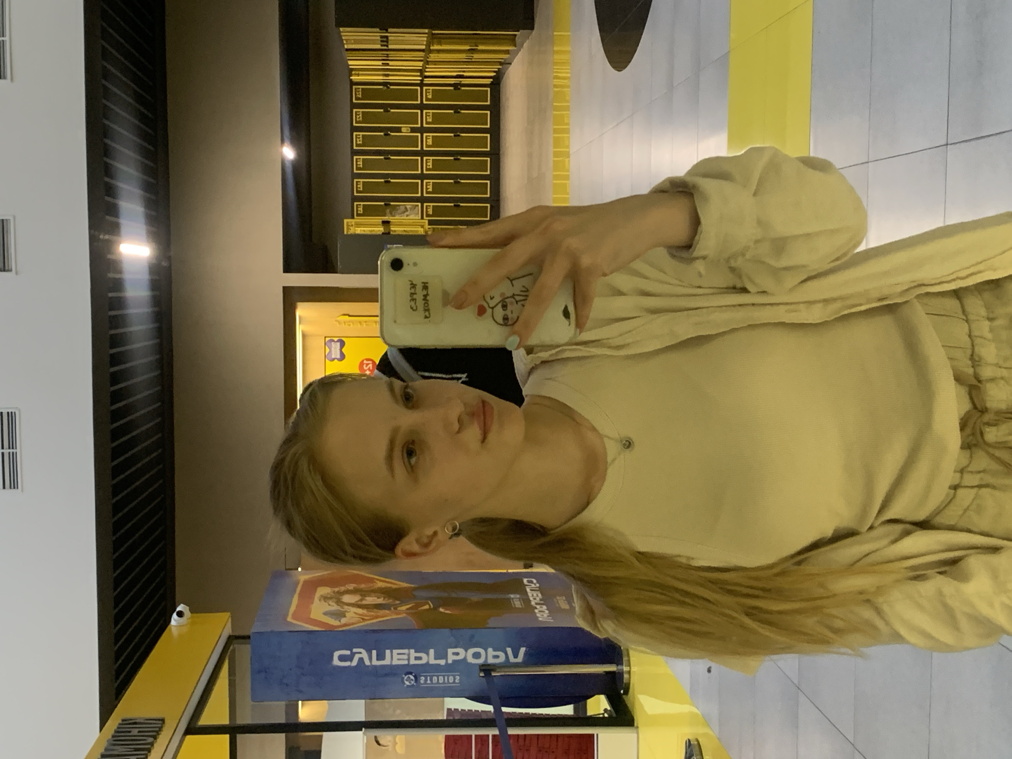 The image depicts a young blonde woman taking a mirror selfie within what appears to be a modern indoor public space in Kyiv, Ukraine. The woman, with her hair in a ponytail, wears a light-colored ribbed top and an open, light-beige long-sleeved jacket. She also wears a small hoop earring in her left ear and a delicate necklace. She holds a white smartphone with a cartoon sticker in her left hand, which has light blue nail polish on her fingernails.
The environment features a gray tiled floor with prominent yellow stripes, leading towards a bank of yellow lockers with vertical black accents on the right side of the frame. In the background, beyond the lockers, more of the tiled floor and a dark, possibly reflective, surface are visible. To the left, a large yellow structure is partially visible, potentially part of a counter or entrance, with a security camera mounted on its underside. Below this yellow structure, a tall blue advertising banner or pillar displays a mirrored image of a fantasy-style character (possibly from a movie poster) and mirrored Cyrillic text, which is indecipherable as a direct location but appears to include "2 STUDIO". The ceiling features a dark grid structure with recessed lighting and air vents.
Based on the distinctive yellow and black color scheme, the modern design, the presence of lockers, and the movie poster on the blue pillar, the location is consistent with the lobby of a "Planeta Kino" cinema, a popular chain in Kyiv, Ukraine. The reflective surfaces and artificial lighting contribute to a clean, commercial aesthetic.