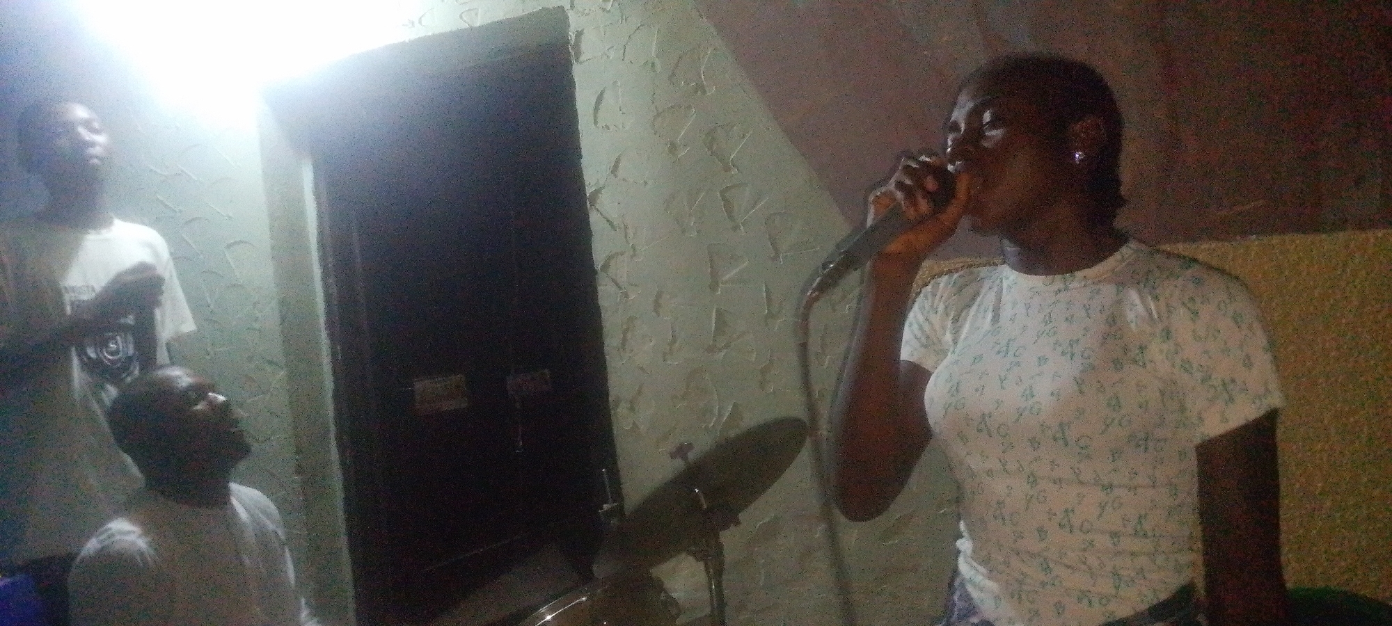 The image shows a dimly lit indoor scene, possibly a private residence or a small performance venue. Three individuals are visible: a woman singing into a microphone on the right, a man seated and looking up on the left, and another man standing behind him. The woman is wearing a patterned t-shirt. The two men are wearing white shirts. A microphone stand and drum set are partially visible. The background includes a textured wall and a closed door. The location is described as being in Lokoja, Nigeria.