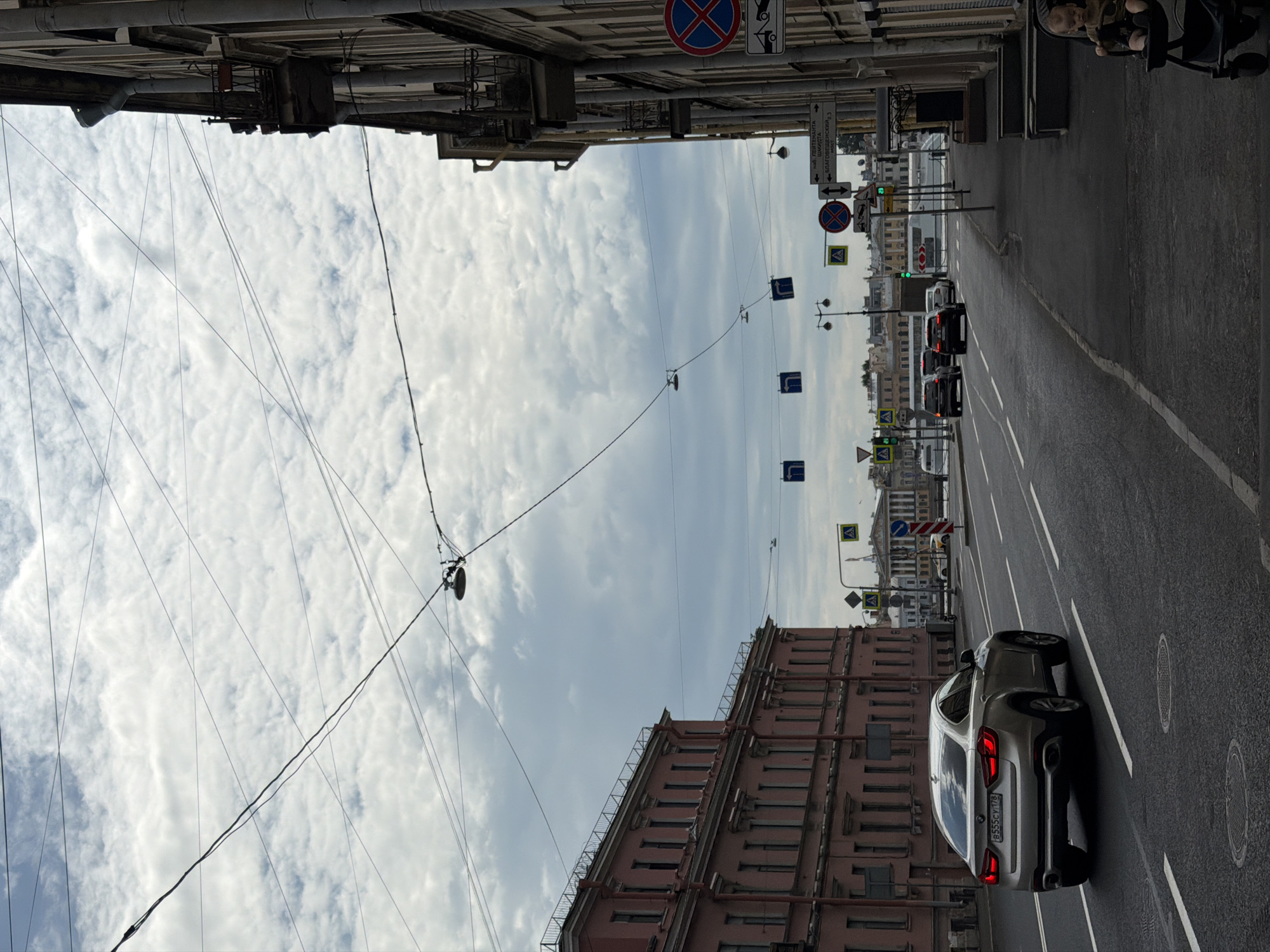 An urban street scene unfolds in Saint Petersburg, Russia, under an overcast sky filled with white, fluffy clouds. Numerous overhead utility wires crisscross the sky above the street.
In the foreground, a silver SUV, resembling a BMW X6, is prominently featured, driving away from the viewer. Its Russian license plate, `B555CY 178`, is clearly visible, with '178' indicating the Saint Petersburg region. Further down the multi-lane street, other vehicles are visible, and a traffic light shows a green signal, suggesting active traffic flow.
The street is lined with historic, multi-story buildings, characteristic of European architecture. On the right side of the street, a series of street signs are mounted. These include blue rectangular signs with white arrows indicating mandatory directions, yellow triangular signs warning of pedestrian crossings, and circular signs like "No Stopping" (a red circle with a blue 'X') and "No Parking" (a red circle with a diagonal line). A large brown directional sign features Cyrillic text, clearly pointing towards "САДОВАЯ" (Sadovaya Street), "НАБ. РЕКИ МОЙКИ" (Moyka River Embankment), and "БЛАГОВЕЩЕНСКИЙ МОСТ" (Blagoveshchensky Bridge), confirming the city's identity.
A person's head and upper body are partially visible in the far upper right corner of the image, possibly in a stroller. The scene captures a typical daytime street environment in Saint Petersburg.