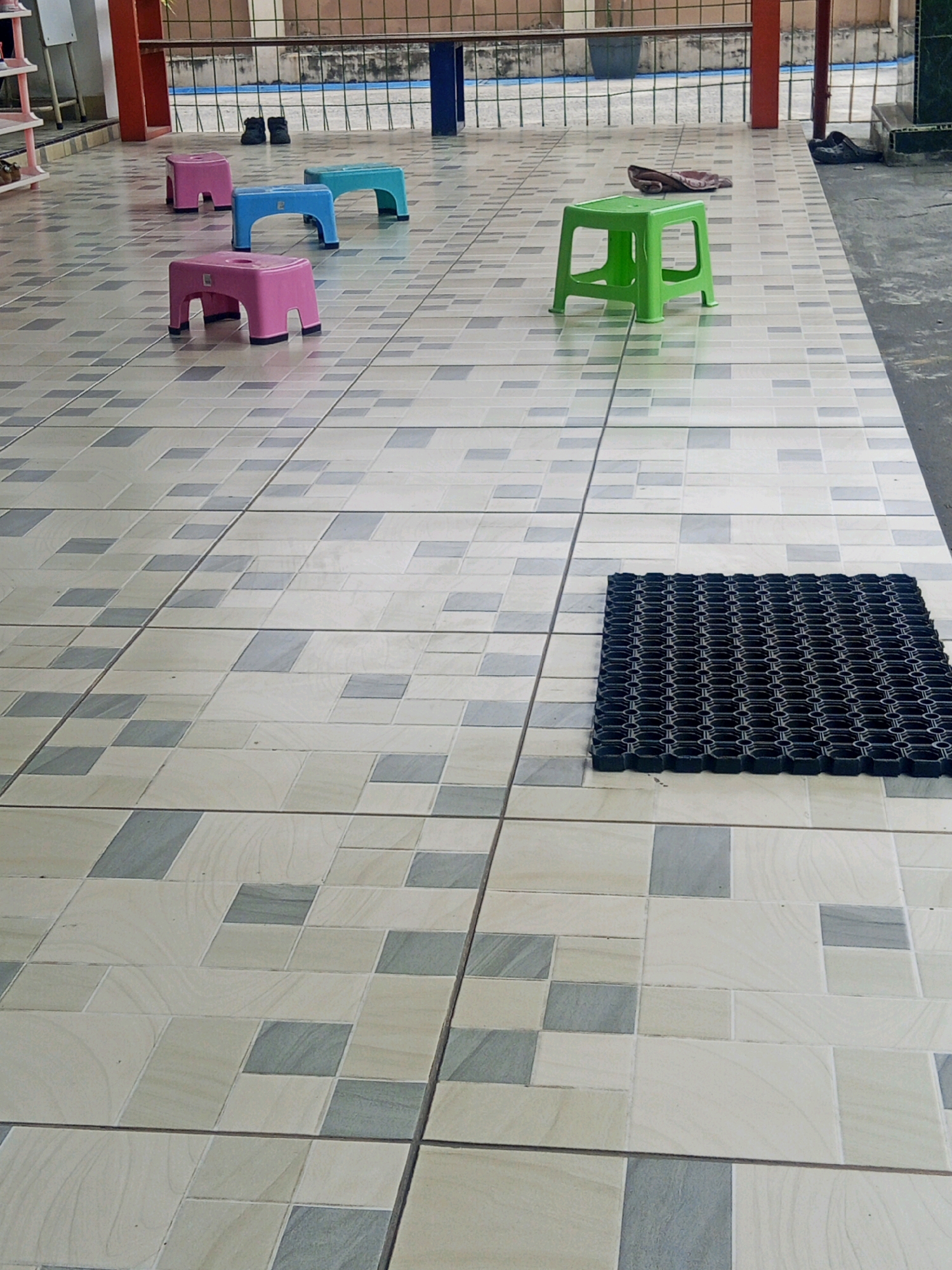 The image shows a tiled outdoor area with several small plastic stools. In the foreground, a black mat with a honeycomb pattern is placed on the tiles. Further back, four stools are arranged: two pink, one blue, and one green. Behind the stools, a metal fence with blue and brown bars is visible, and beyond that, a blurry background that appears to be a building or walkway. The tiling on the floor is a pattern of light-colored squares with some darker grey and light beige rectangular tiles interspersed. The overall impression is of a public outdoor space, possibly a waiting area or a children's play area. The location in Lubuklinggau, Indonesia, suggests a warm climate. There are no people visible in the image.