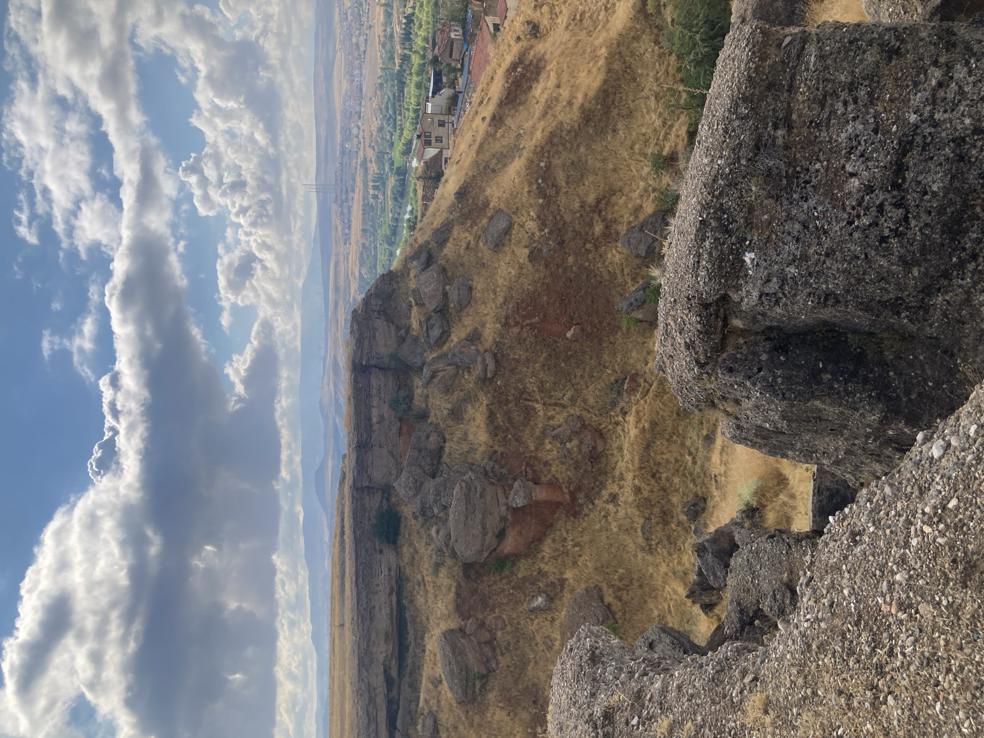 This image captures a panoramic view from a rocky overlook in Avanos İlçe Merkezi, Turkey. The foreground is dominated by large, rough-textured rocks and gravel, suggesting a natural or perhaps archaeological site. Beyond these rocks, a steep, dry, grassy slope descends, dotted with smaller boulders and patches of reddish earth. In the middle ground, a small town is visible, characterized by houses with red tile roofs, nestled amongst greenery and alongside a winding river. The background reveals a vast, arid landscape with rolling hills and distant mountains, under a dynamic sky filled with dramatic clouds. Three tall towers, possibly radio or communication masts, rise from the town in the distance. The scene appears to be a sunny day, indicated by the bright sky and the way the light illuminates the landscape. There are no people or specific actions depicted, only the static environment.