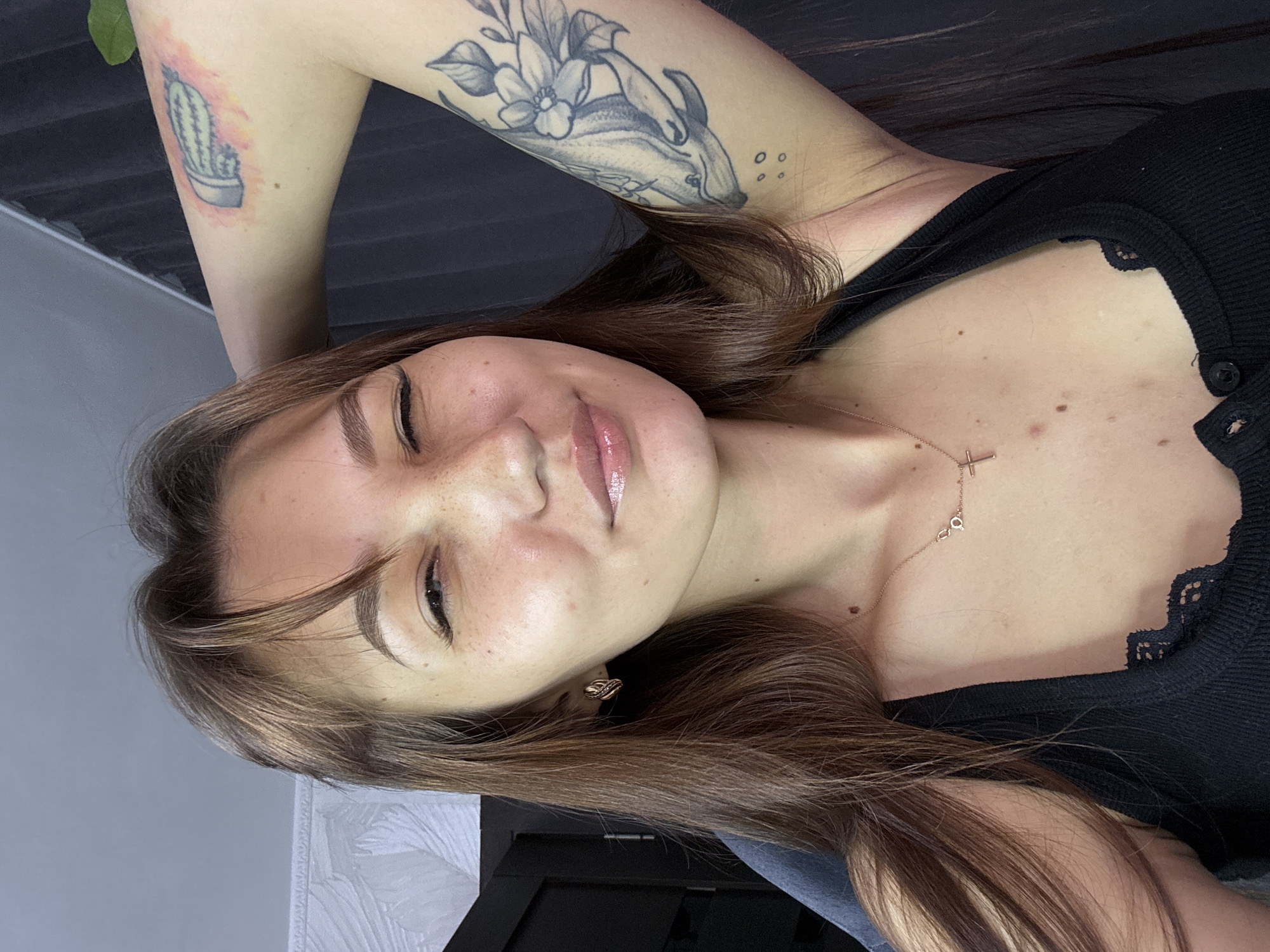 A young woman with brown hair and freckles is shown in a close-up, rotated portrait. She is wearing a black top with lace trim and a delicate cross necklace. Her arm is visible, featuring a tattoo of a cactus on her upper arm and a floral design on her forearm. Her expression is soft, with her eyes partially closed and her lips slightly parted, suggesting a relaxed or sensual mood. The background is a mix of dark wood and a light gray textured surface, possibly an indoor setting. The image has a warm, intimate feel. The time of day and weather are not discernible from the image.