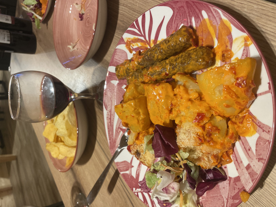 The scene depicts a meal served on a wooden table in an indoor setting in the city of Palež, Montenegro. In the foreground, a white plate with a red/pink leaf pattern holds a main course consisting of chunks of potatoes coated in a thick, bright orange-red sauce, two cylindrical meat items also covered in the same sauce, and a side portion of mixed green salad with shredded vegetables. A silver fork rests on the plate. To the left, a white bowl with a red rim contains plain potato chips. Centered in the midground is a wine glass containing red wine, approximately one-quarter full. To the right of the wine glass, an empty white bowl with a red rim and leaf pattern, matching the main plate, contains minimal salad remnants. In the blurry background, dark-colored bottles and light-toned furniture are faintly visible. No people are present.