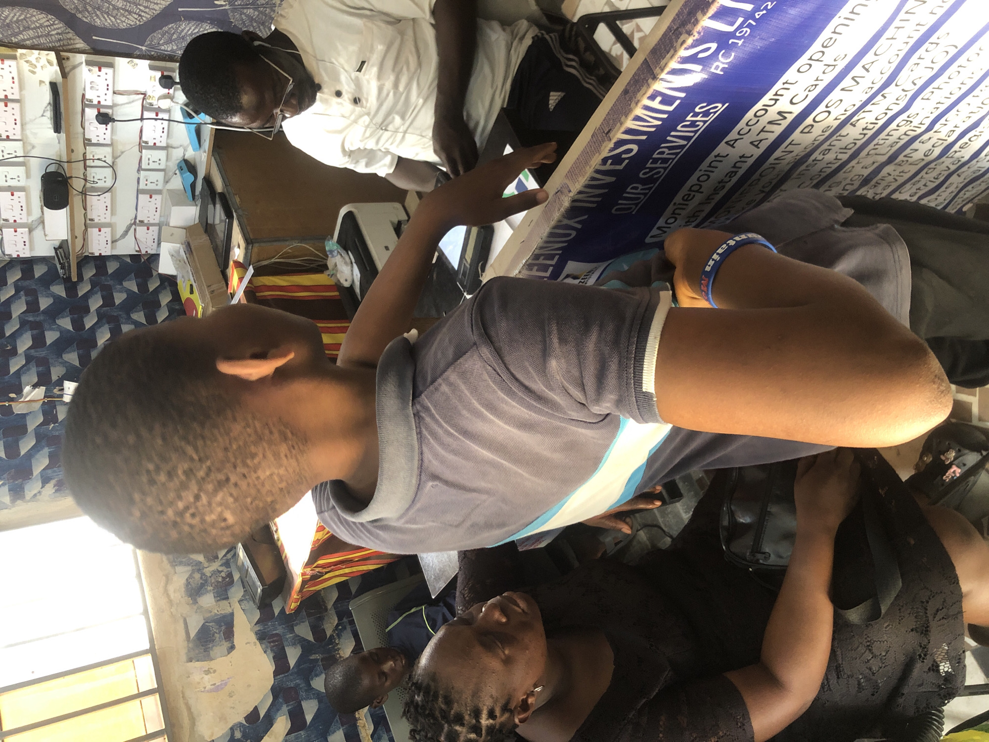 In Ado, Nigeria, an indoor scene captures a transaction or interaction at what appears to be a small business. In the foreground, a young boy, seen from the back, wears a grey polo shirt with blue and white stripes on the collar and sleeves. His right arm is extended towards a blue sign with white text that reads, "WEENUX INVESTMENTS LTD OUR SERVICES" and "Moniepoint Account opening." He wears a blue wristband with white lettering.
To the left, a woman wearing a black lace dress and earrings looks towards the boy. She has dark skin and braided hair. A black handbag rests on her lap, and a strap hangs down her arm.
In the background, a man with glasses and a white polo shirt sits at a desk, looking down at something in front of him. He wears earphones and has dark skin. Behind him, a wall is covered with numerous white electrical outlets and switches. Various items, including white boxes, a black speaker, and a printer, are visible on shelves and surfaces. Another young boy in a blue shirt is seated in a chair to the left of the man. The overall environment suggests a busy, functional space with a focus on financial services or business operations.